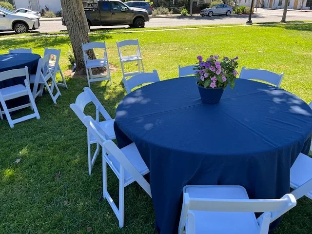 Round table covered with a blue tablecloth surrounded by white garden chairs.