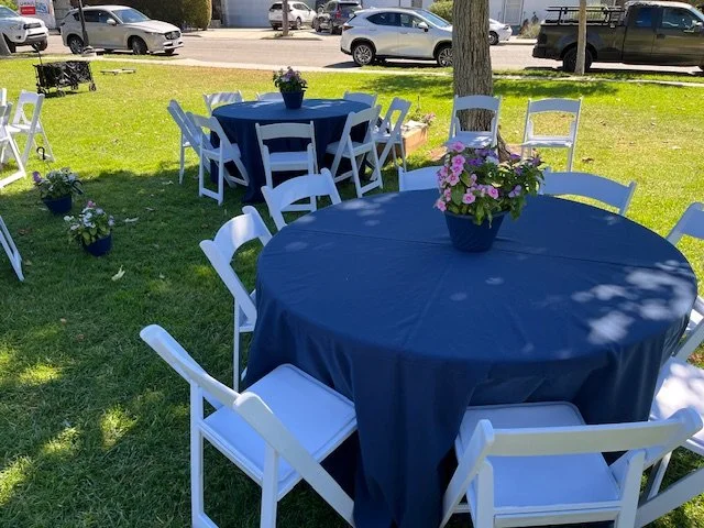 Round table covered with a blue tablecloth surrounded by white garden chairs.