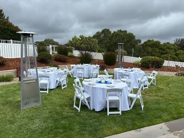 Outdoor backyard setup with round tables covered in white tablecloths, surrounded by white folding chairs, and topped with tableware, in front of a white fence and landscaped plants with a house and cloudy sky in the background.