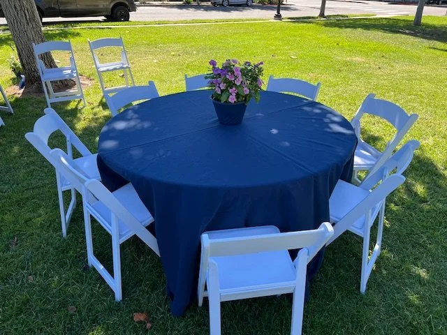 Round table covered with a blue tablecloth surrounded by white garden chairs.