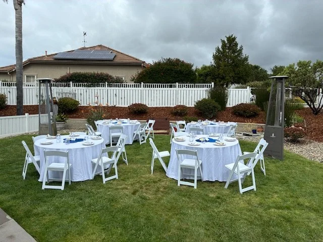 Outdoor backyard setup with round tables covered in white tablecloths, surrounded by white folding chairs, and topped with tableware, in front of a white fence and landscaped plants with a house and cloudy sky in the background.