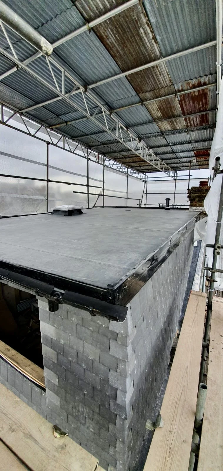 Construction site with a building roof under construction, showing a brick chimney on a log foundation with scaffolding around and a metal roof above.
