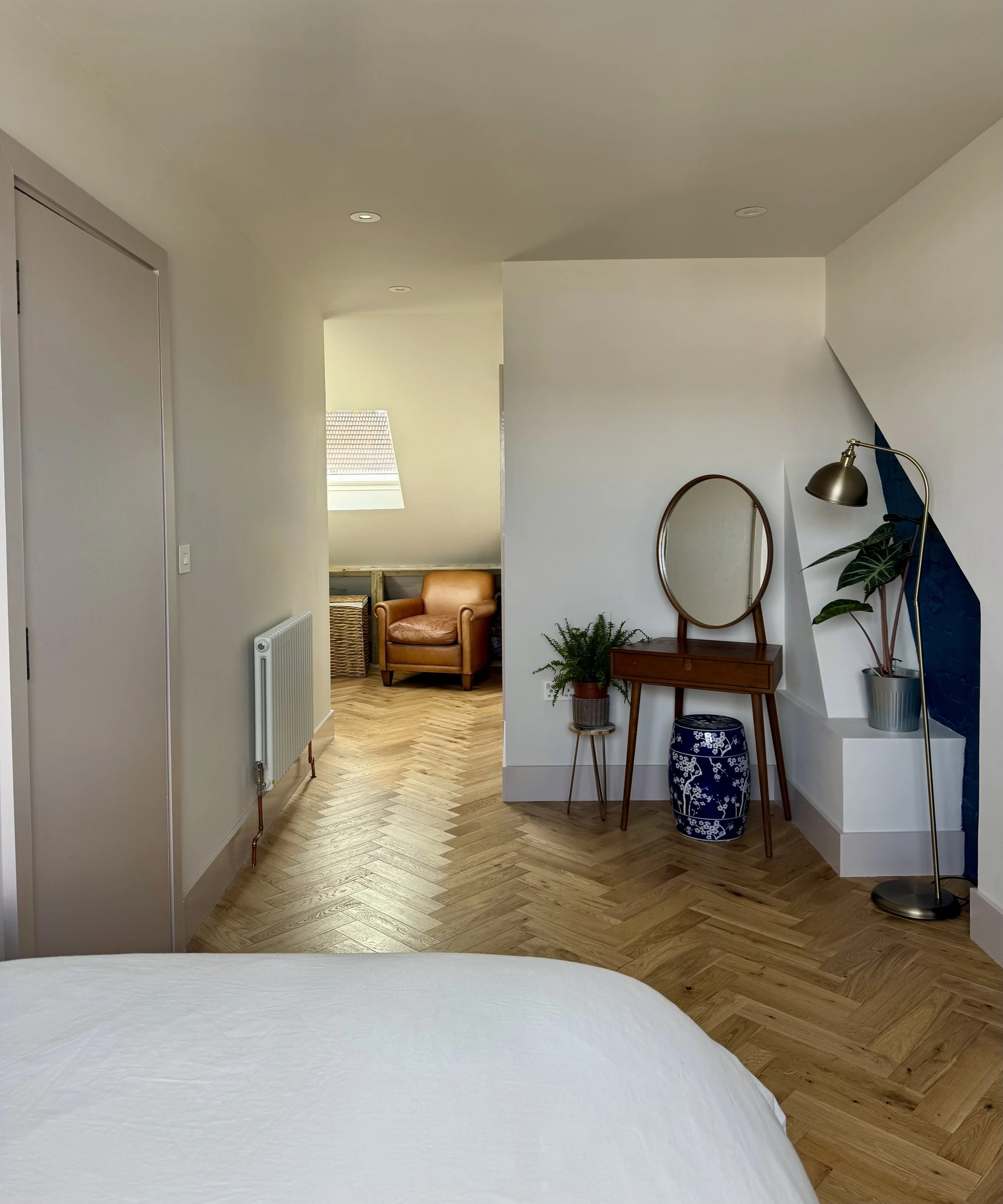 Modern bedroom with hardwood floors, a corner vanity with a mirror, a floor lamp, and potted plants. A sitting area with a leather armchair and a window with a sloped ceiling in the background.