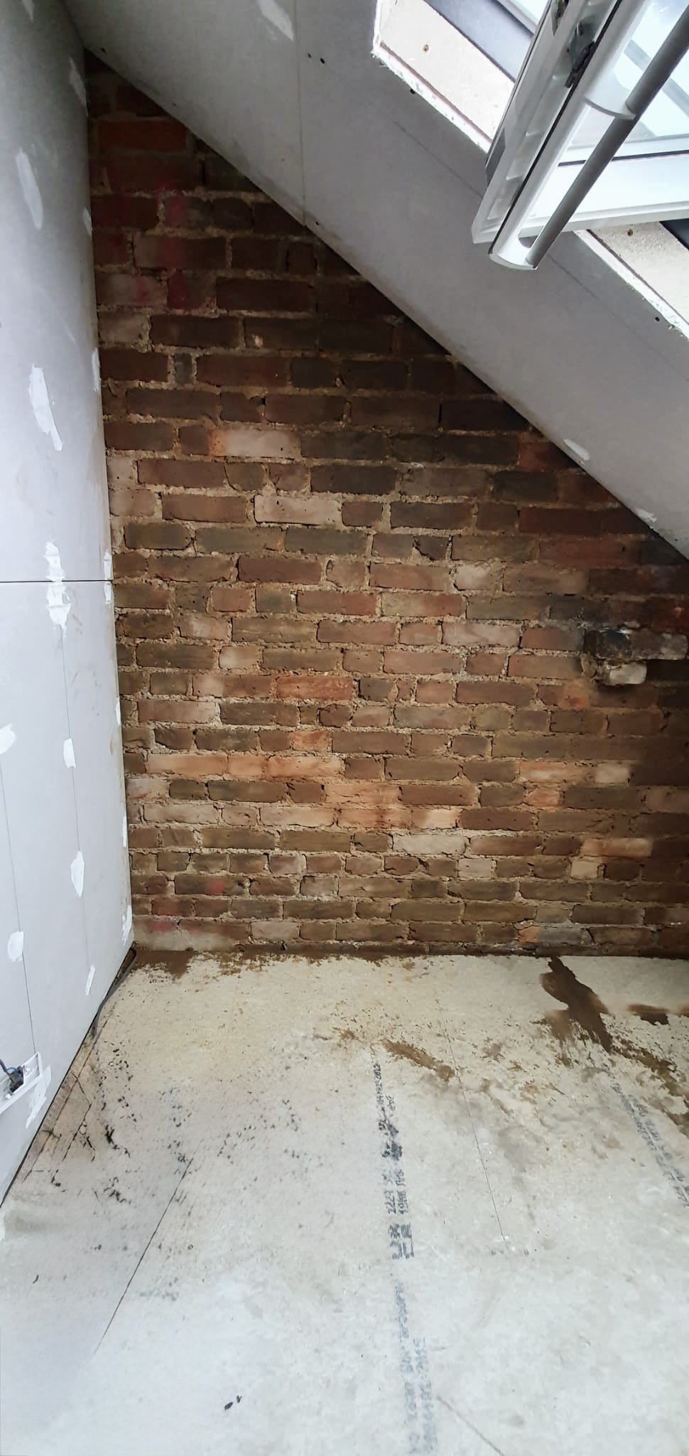 Unfinished indoor space with exposed brick wall, partially installed drywall, and a window with open blinds.