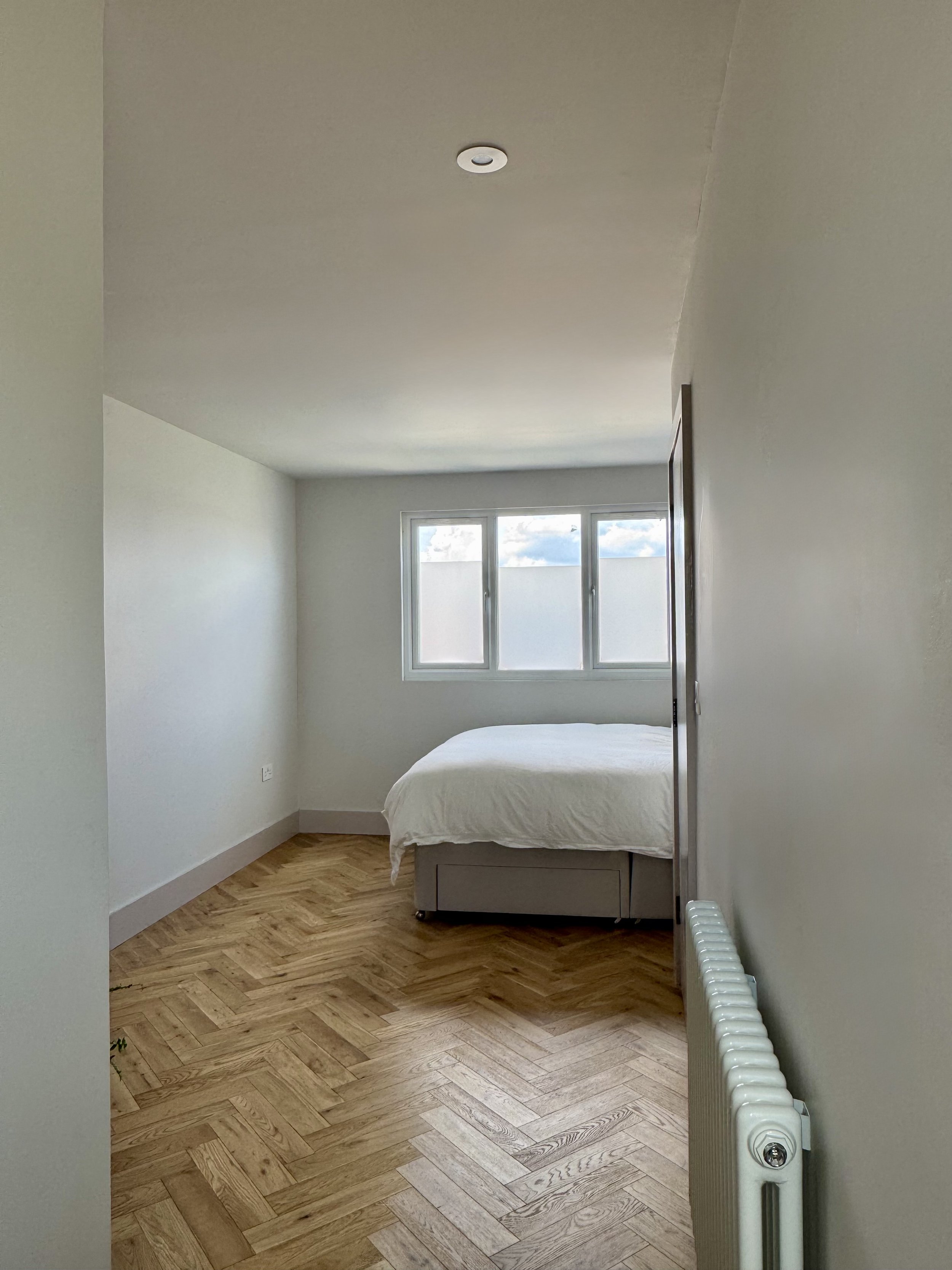 A minimalistic bedroom with a bed, wooden parquet floor, a large window, and plain white walls.