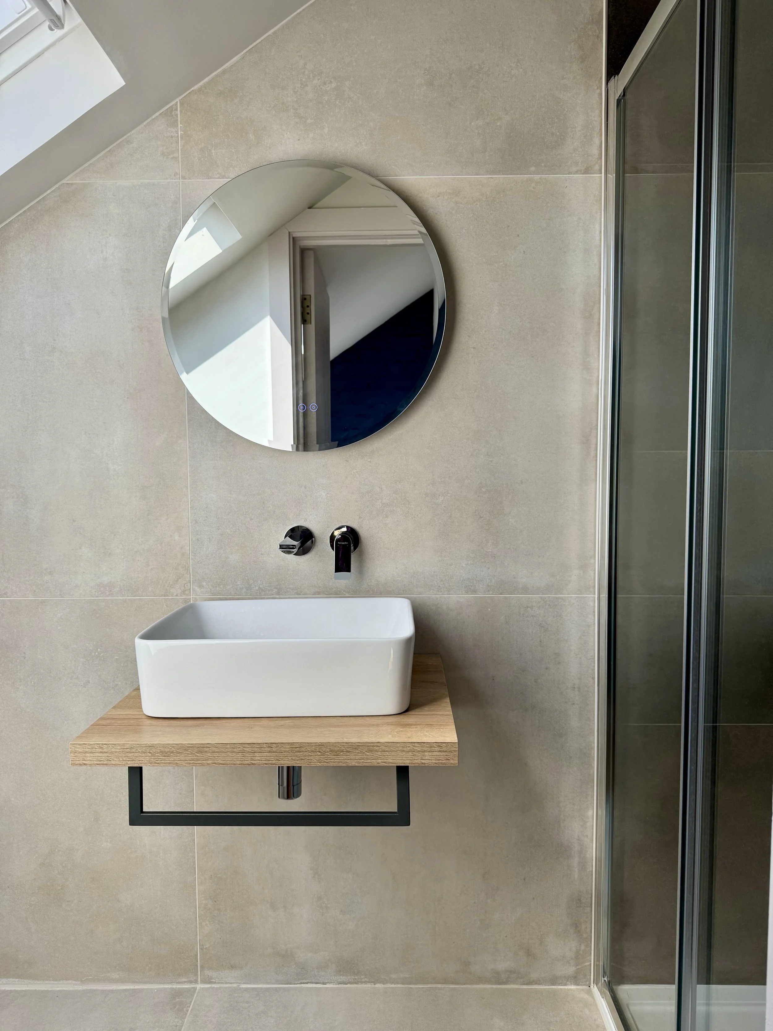 Modern bathroom with a white rectangular vessel sink on a wooden vanity, a round wall mirror, beige tiled wall, and a glass shower enclosure.