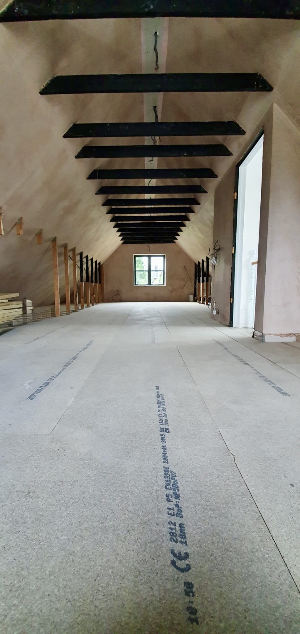 An unfinished attic room with a sloped ceiling, black ceiling beams, and a window at the far end. Wooden framework and construction materials are visible along the sides.