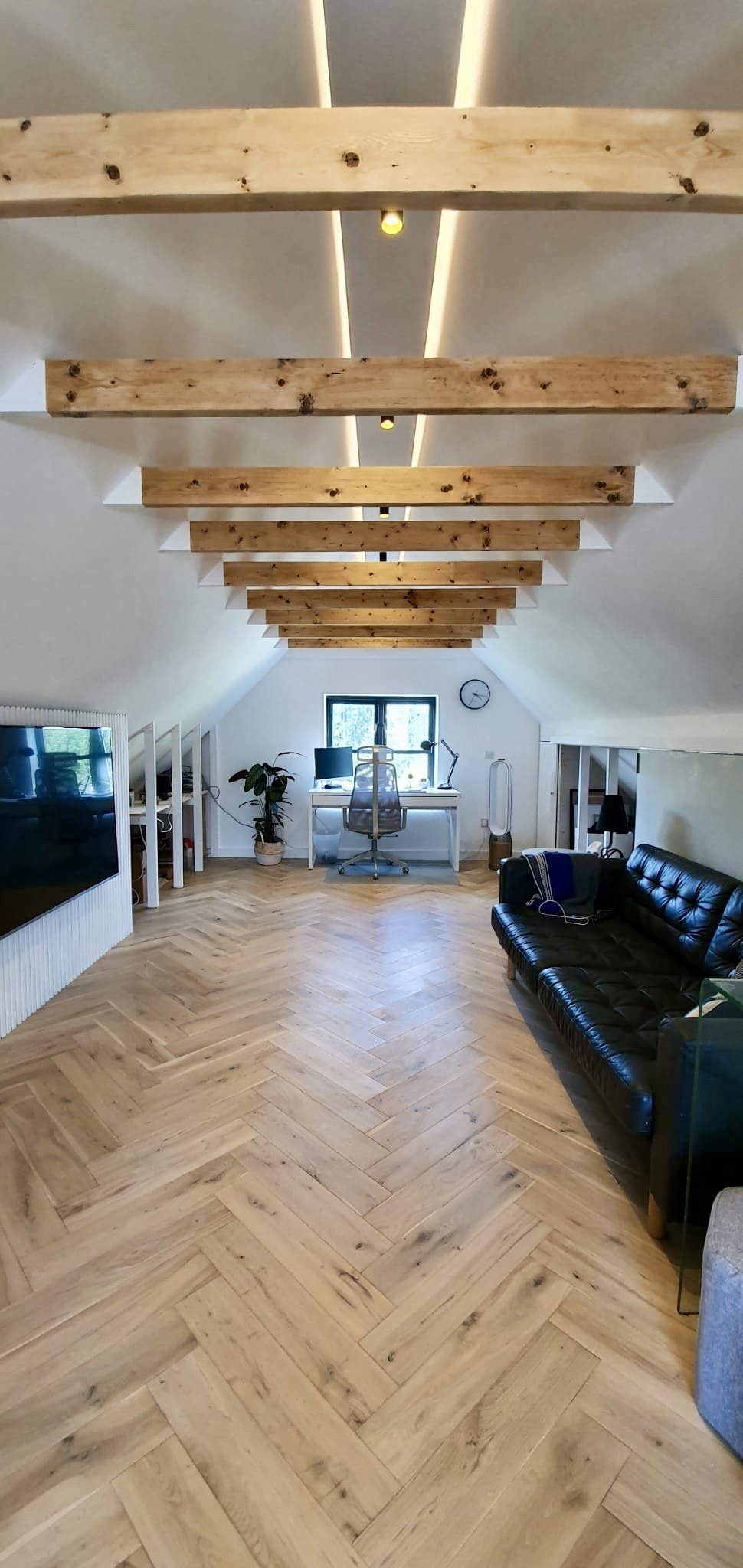 A spacious attic room with a sloped ceiling featuring exposed wooden beams and modern lighting. The room has a wooden herringbone floor, a black leather sofa on the right, a white desk with a chair and computer near the window, and a potted plant to 