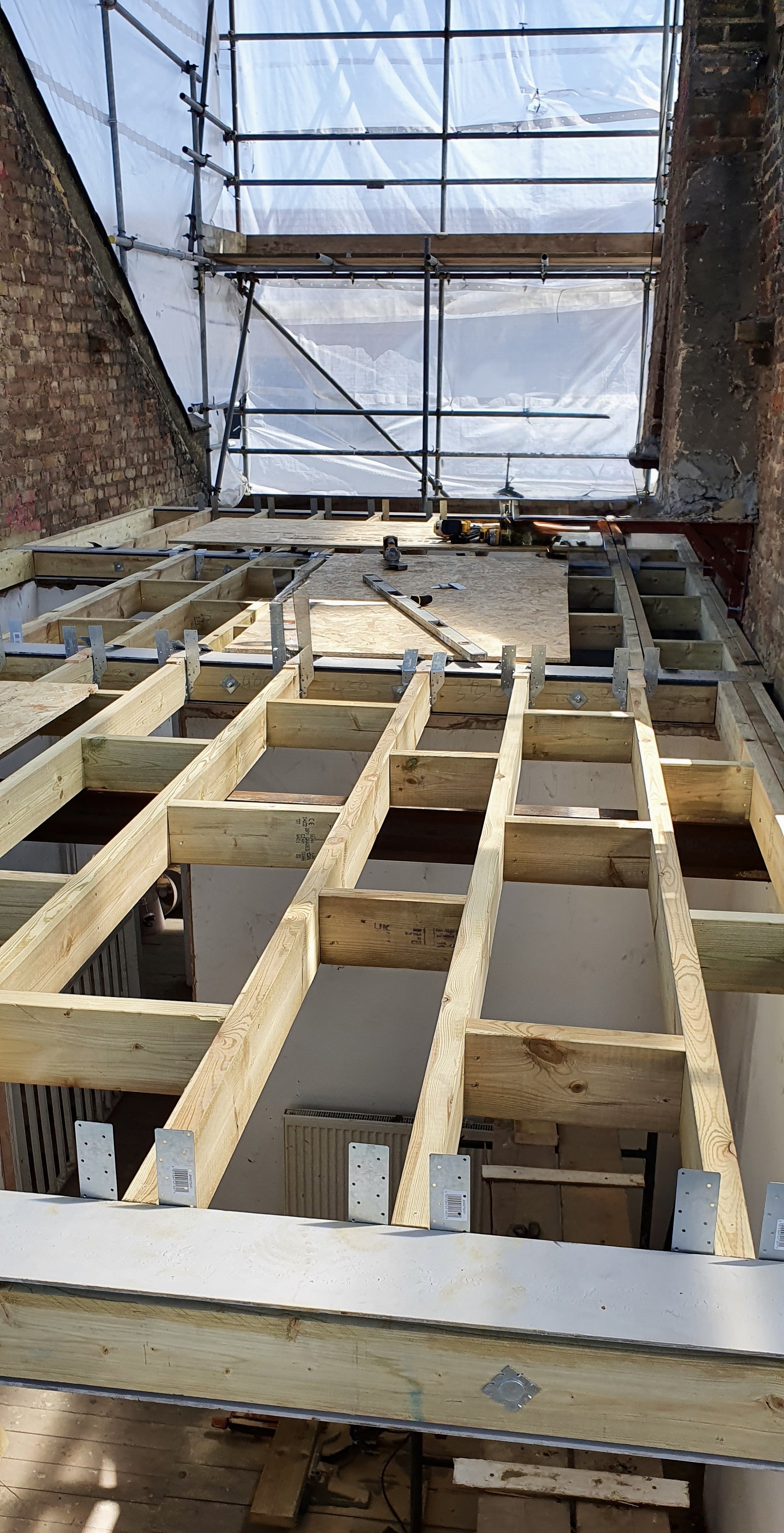 Interior view of a construction site showing wooden floor joists and scaffolding, with plastic sheeting covering part of the space.