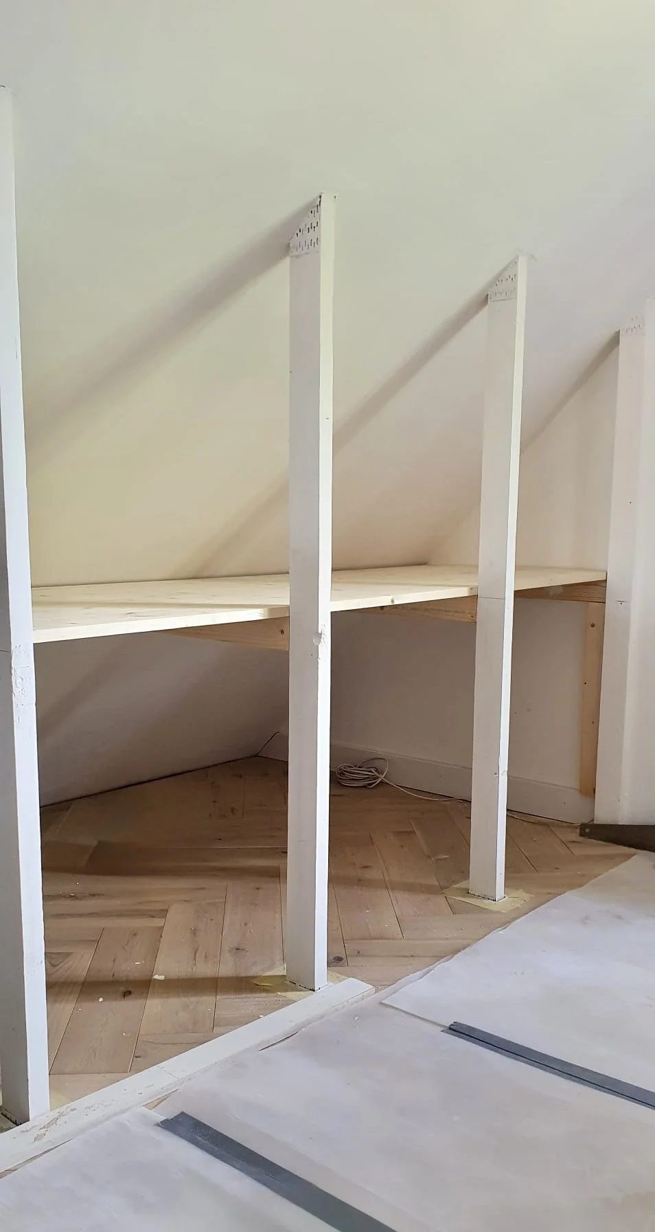Unfinished built-in wooden bookshelf with vertical supports and a sloped back wall in a room under construction.
