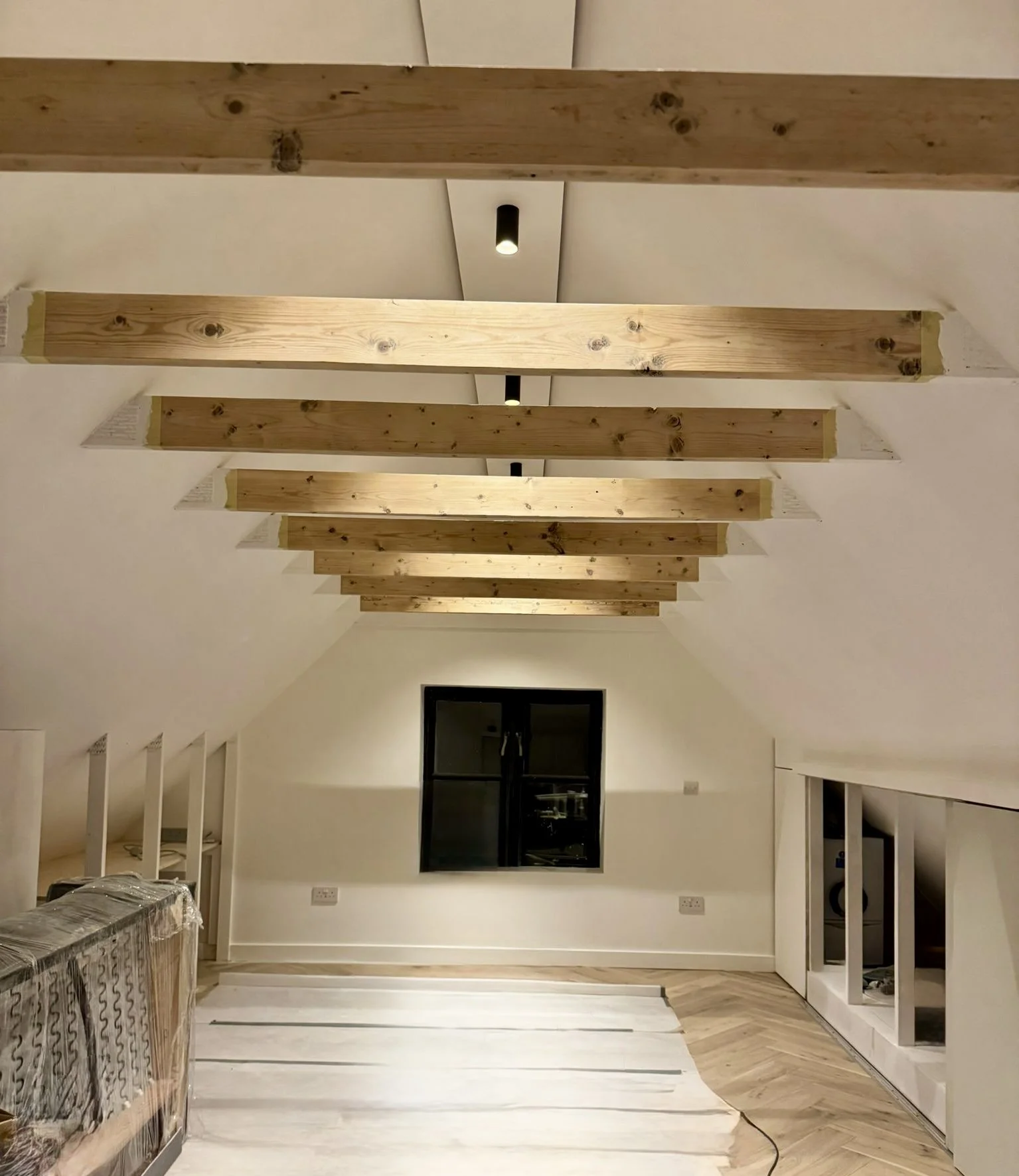 Interior room with sloped ceiling, exposed wooden beams, black window, and partially finished walls.