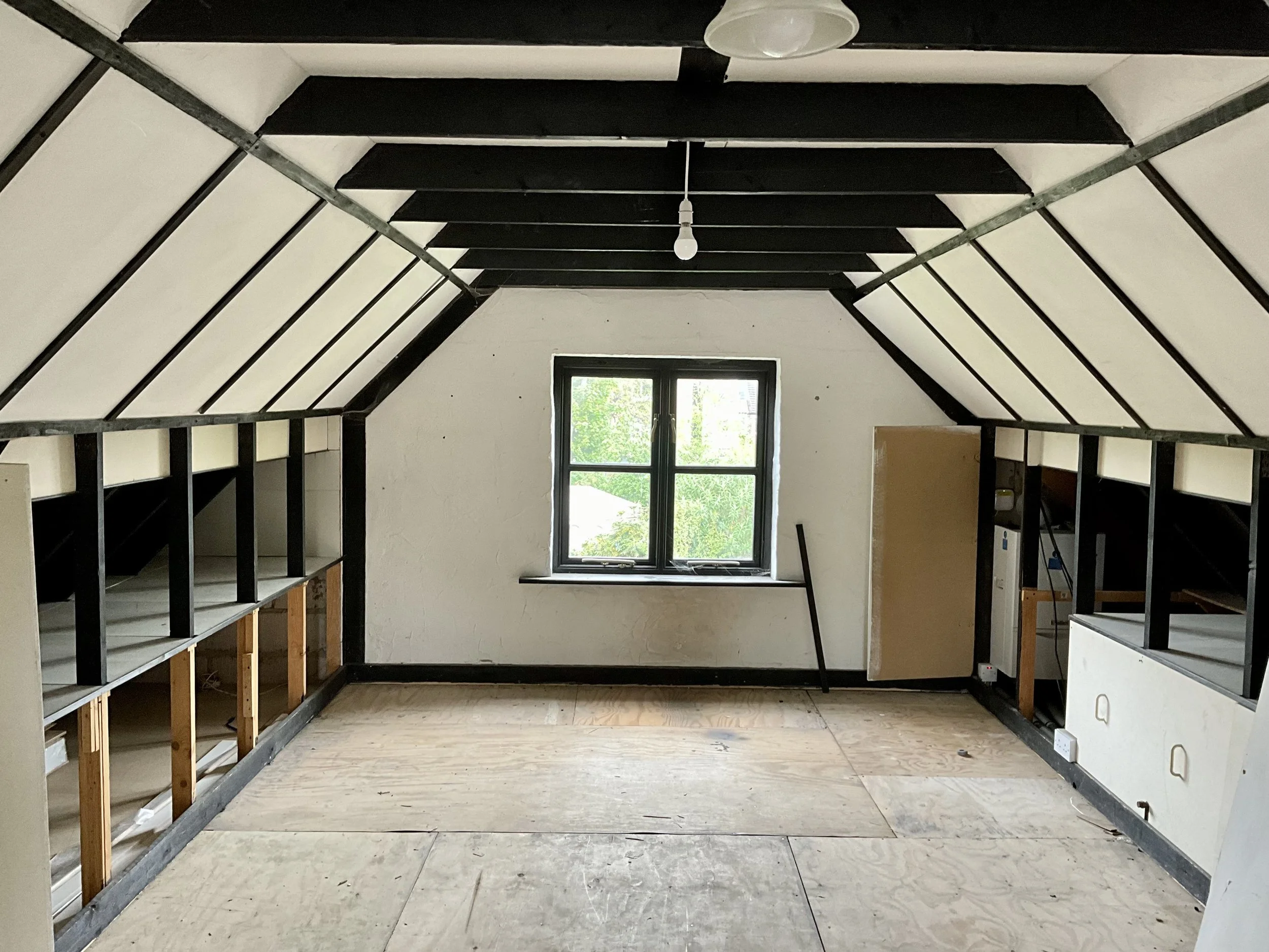 A room under renovation with an angled ceiling, a window in the center, and exposed framing on the sides. The floor is unfinished with plywood sheets, and there are black-painted beams and trim along the walls and ceiling.