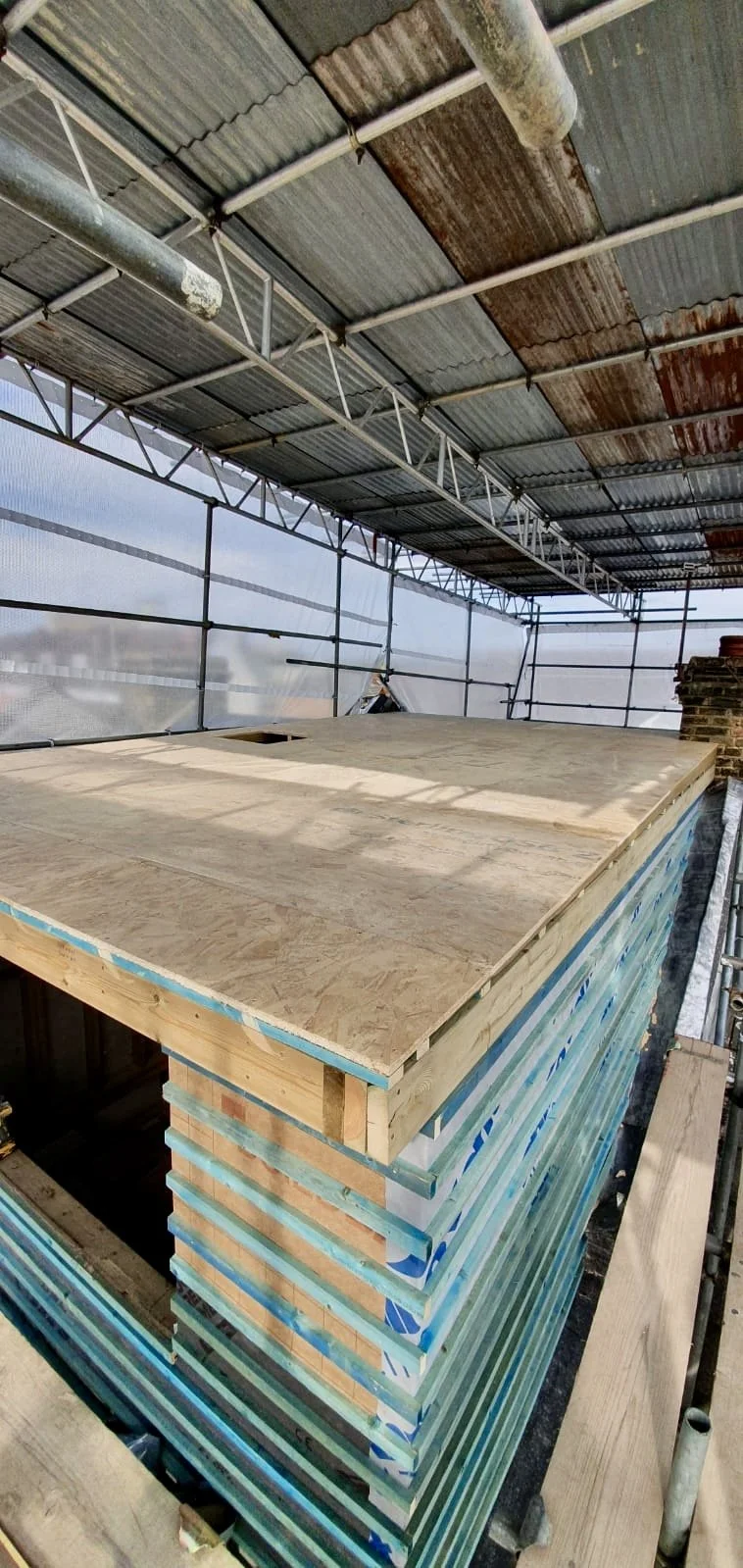 Construction site on a building deck with plywood sheets and stacked framing materials, surrounded by scaffolding and metal roofing.