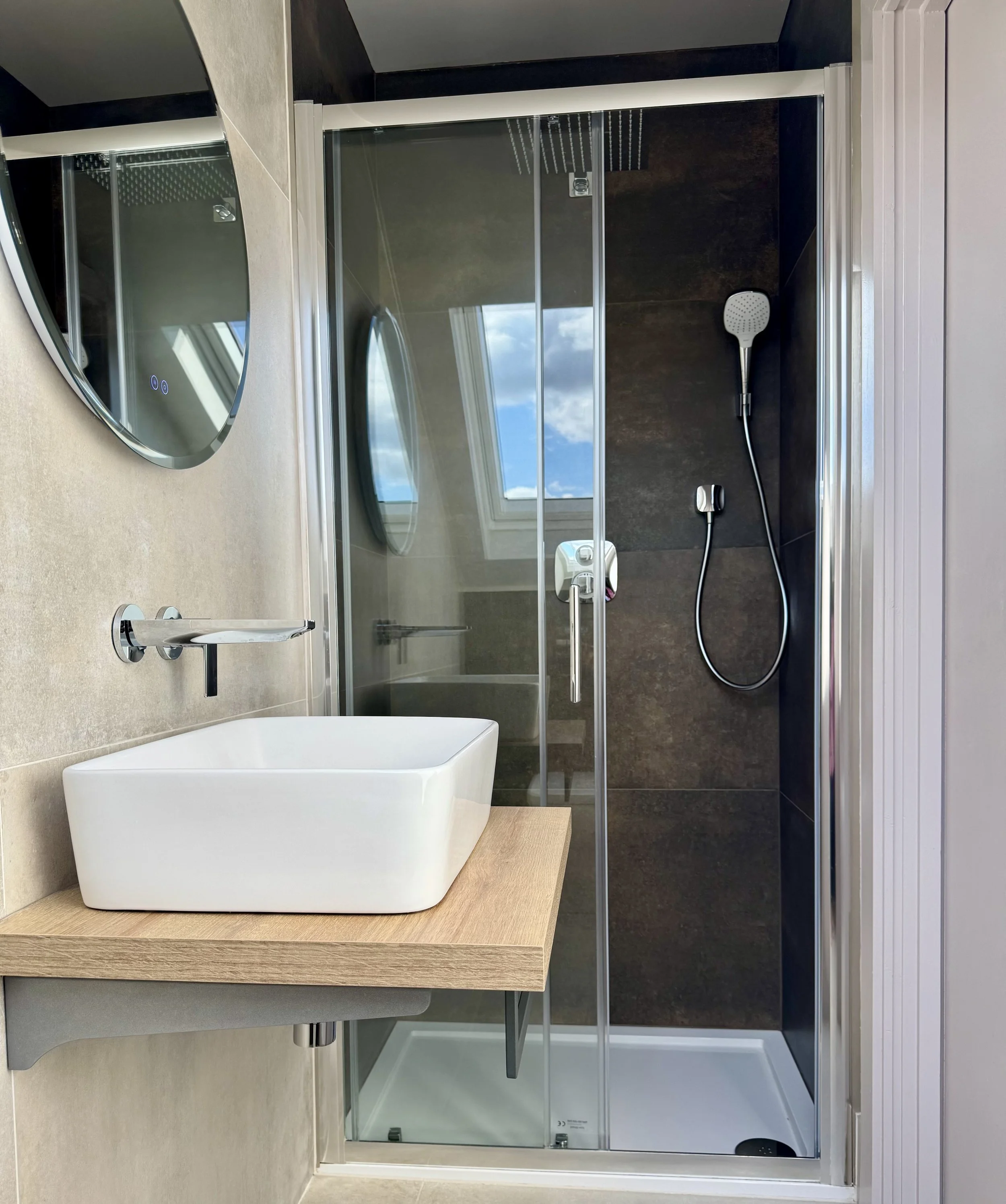 Modern bathroom with a glass shower, white vessel sink on a wooden countertop, round mirror, and a window showing blue sky and clouds.