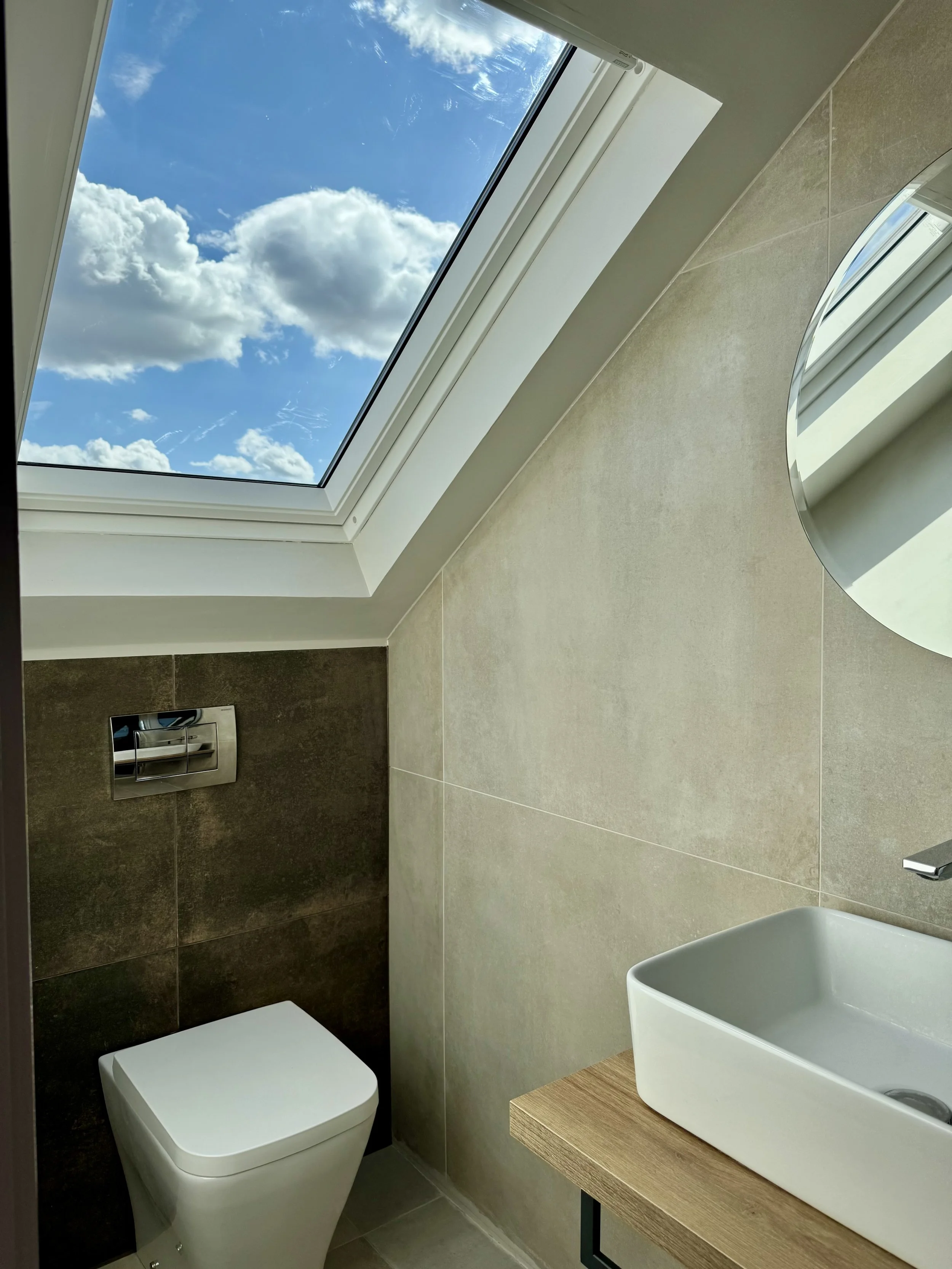 Modern bathroom with a skylight showing blue sky and clouds, a white toilet, a wooden vanity with a square white sink, and a circular mirror.