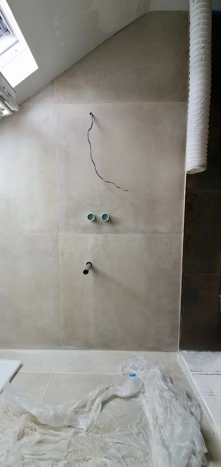 Bathroom wall with pipes and wiring for a sink installation, plastic sheet on the floor, and partially visible window.