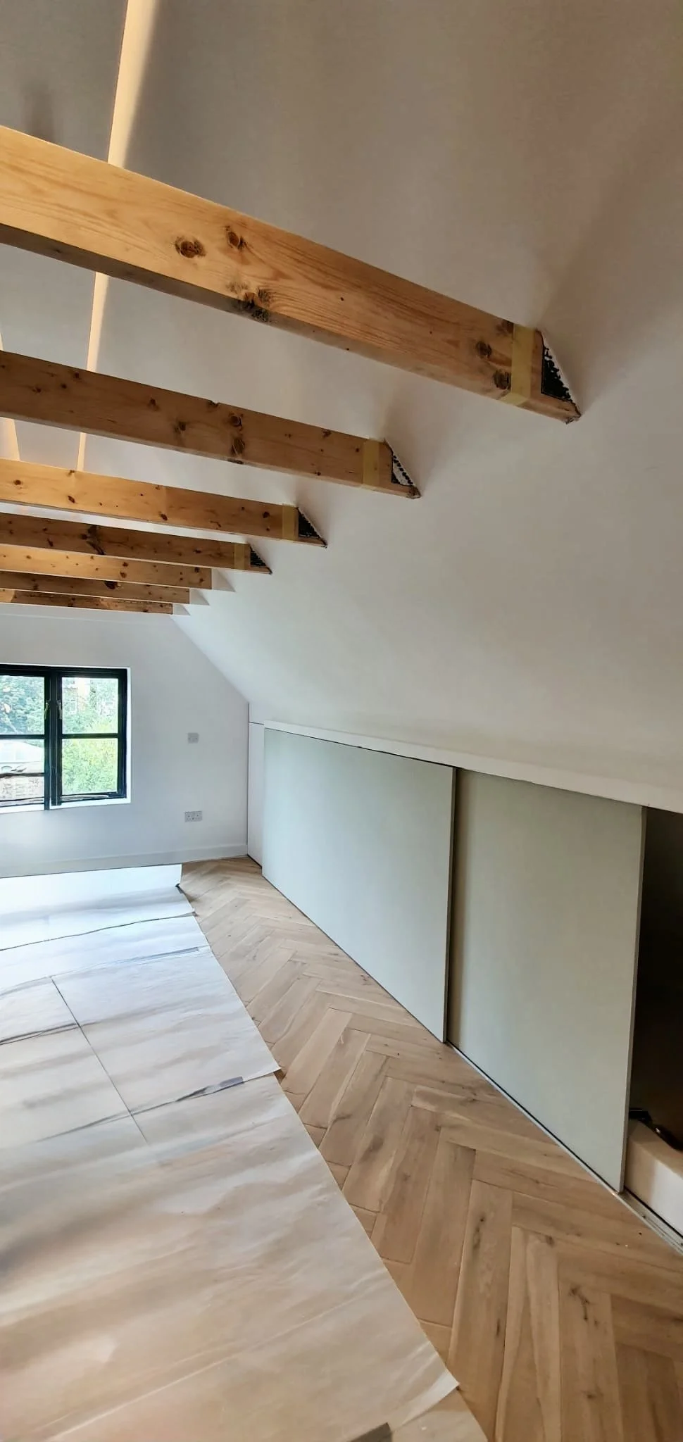Interior of a room under construction with wooden beams on the ceiling, a window on the wall, and partially installed flooring.