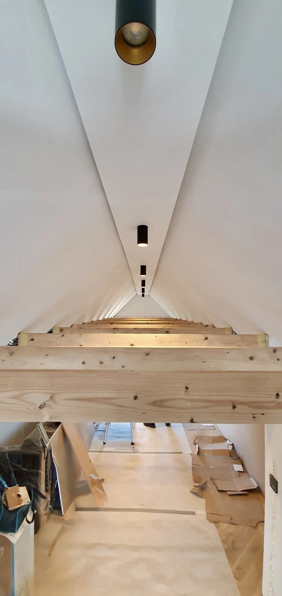 Inside a building under construction, a staircase with wooden steps is being built, viewed from the bottom looking up. The ceiling has black cylindrical light fixtures, and there are construction materials and tools scattered around.