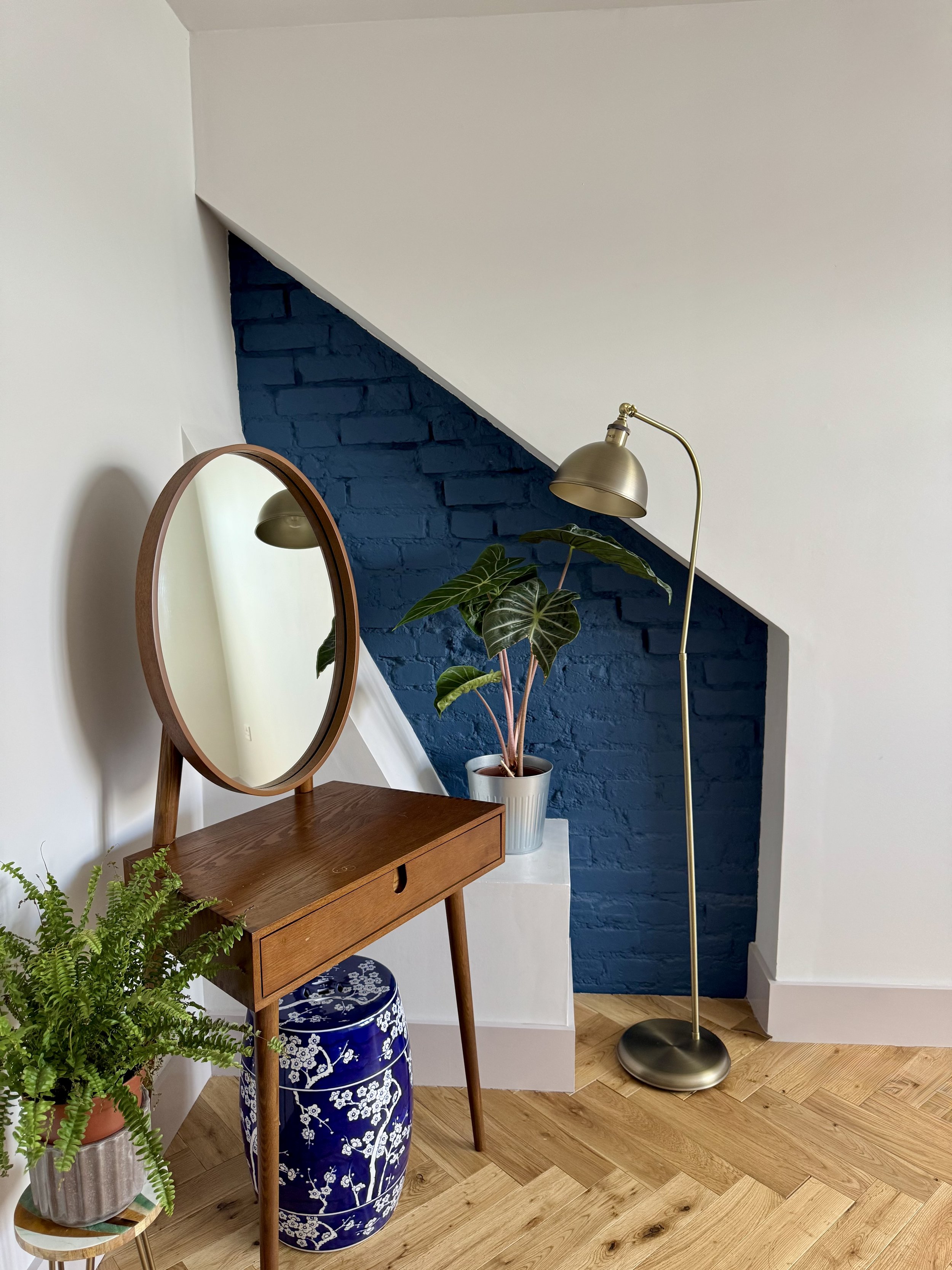 A wooden vanity table with an oval mirror, a potted plant with large green leaves, a blue and white ceramic stool, and a brass floor lamp in front of a blue brick accent wall under a staircase.