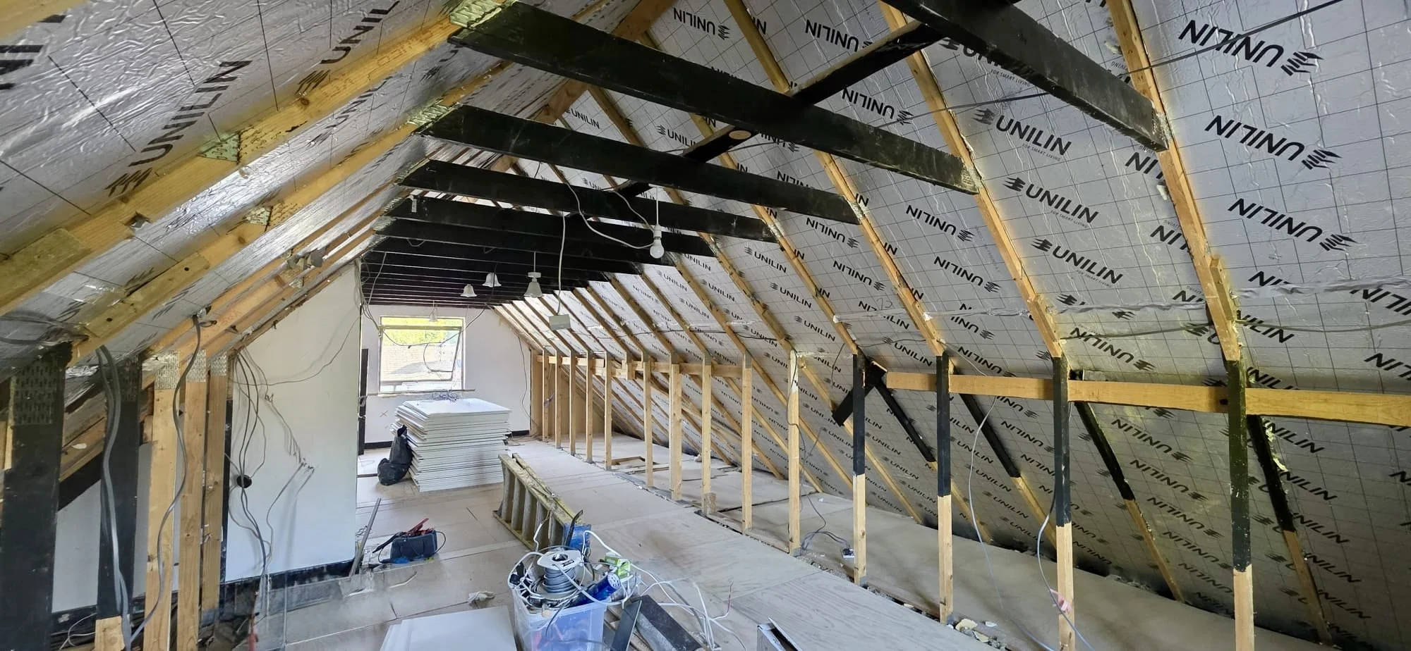 Attic under renovation with exposed wooden beams, insulation, hanging light bulbs, construction tools, and materials.