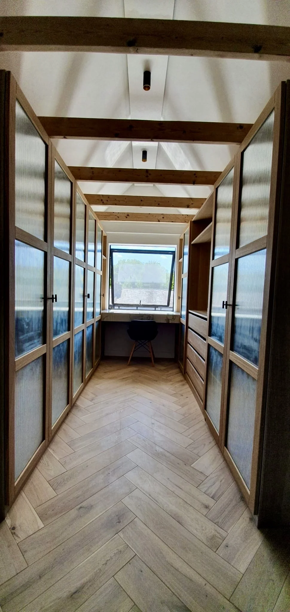 A narrow indoor hallway with wooden slatted upper walls, glass-paneled cabinets on both sides, a small window at the end with a desk and chair underneath, and a white ceiling with exposed wooden beams.