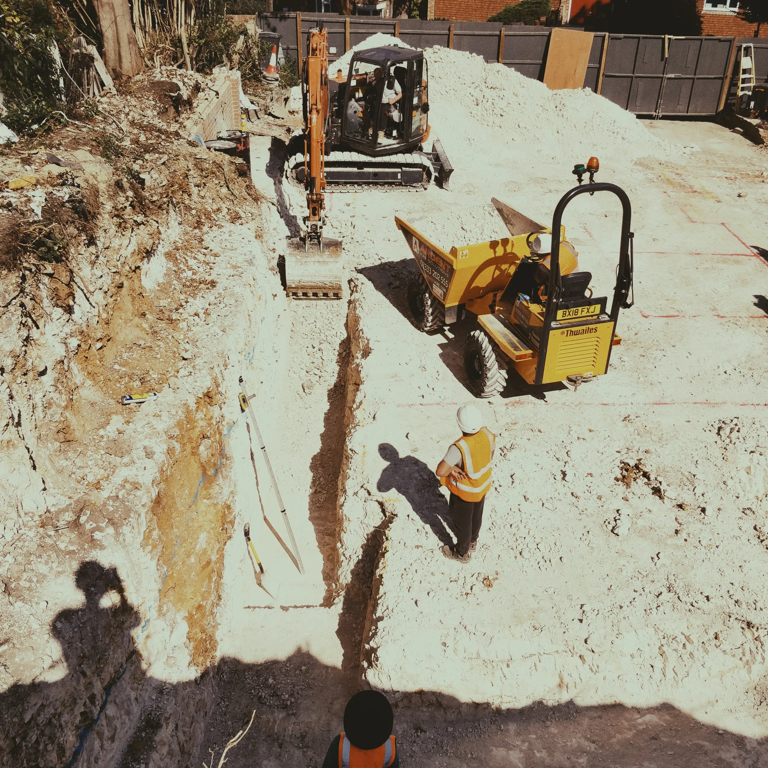 Construction site with two workers wearing yellow safety vests and white hard hats. One worker is looking at a deep trench, and the other is standing nearby. An excavator and a compact roller are present on the site, which is surrounded by a fence an