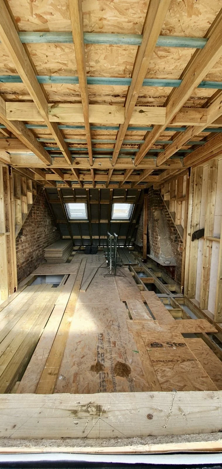 Interior view of an attic under construction with exposed wooden framing, plywood subfloor, and skylight windows.