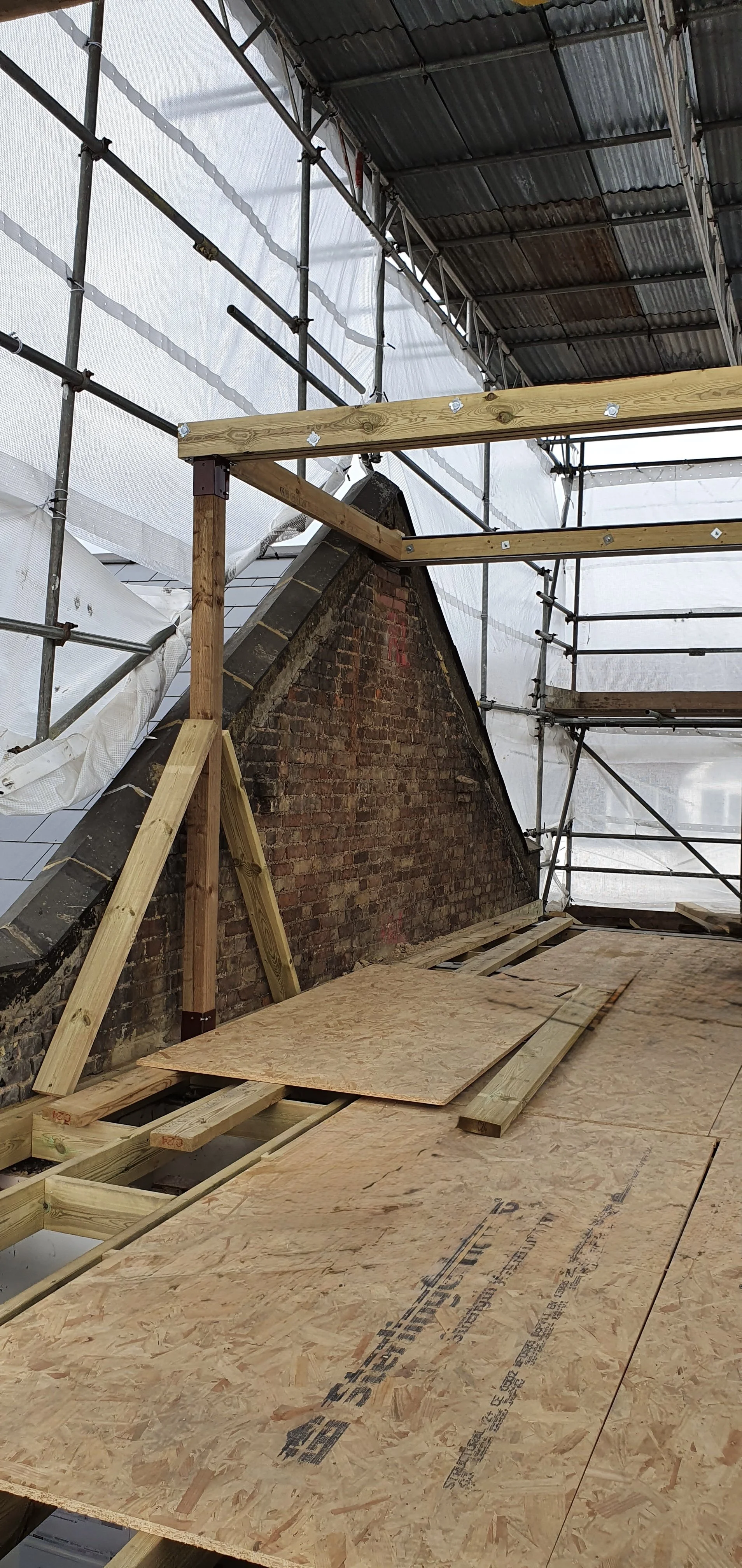 Under construction with plywood floor, wooden beams, and scaffolding surrounding a brick chimney inside a building renovation project.