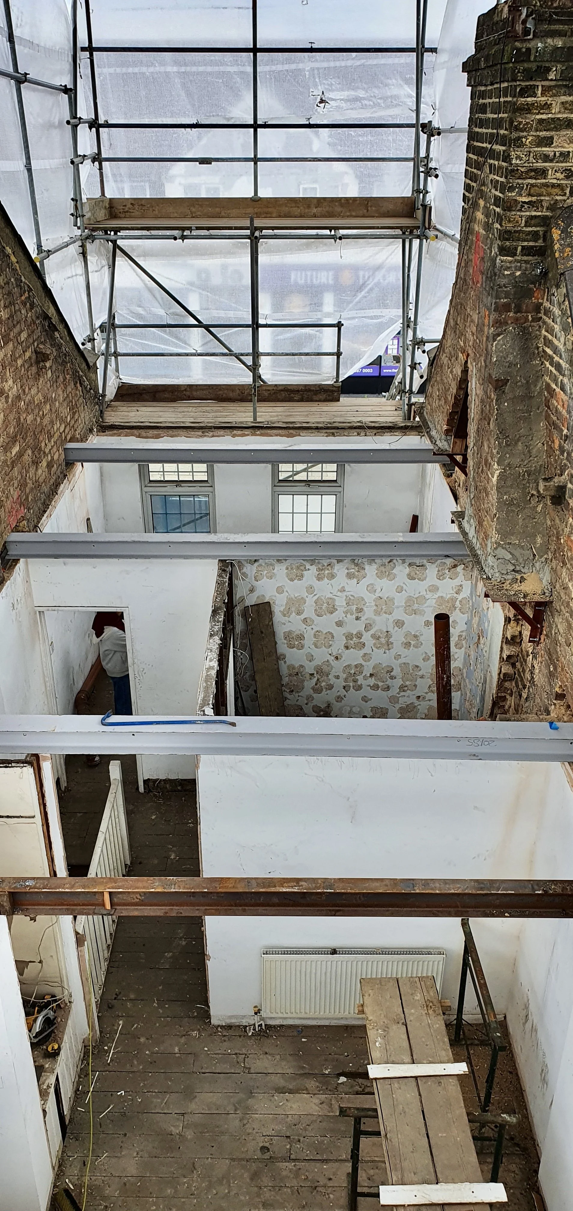 Interior view of a building under renovation, with exposed brick and plaster walls, wooden and metal scaffolding, and construction materials.