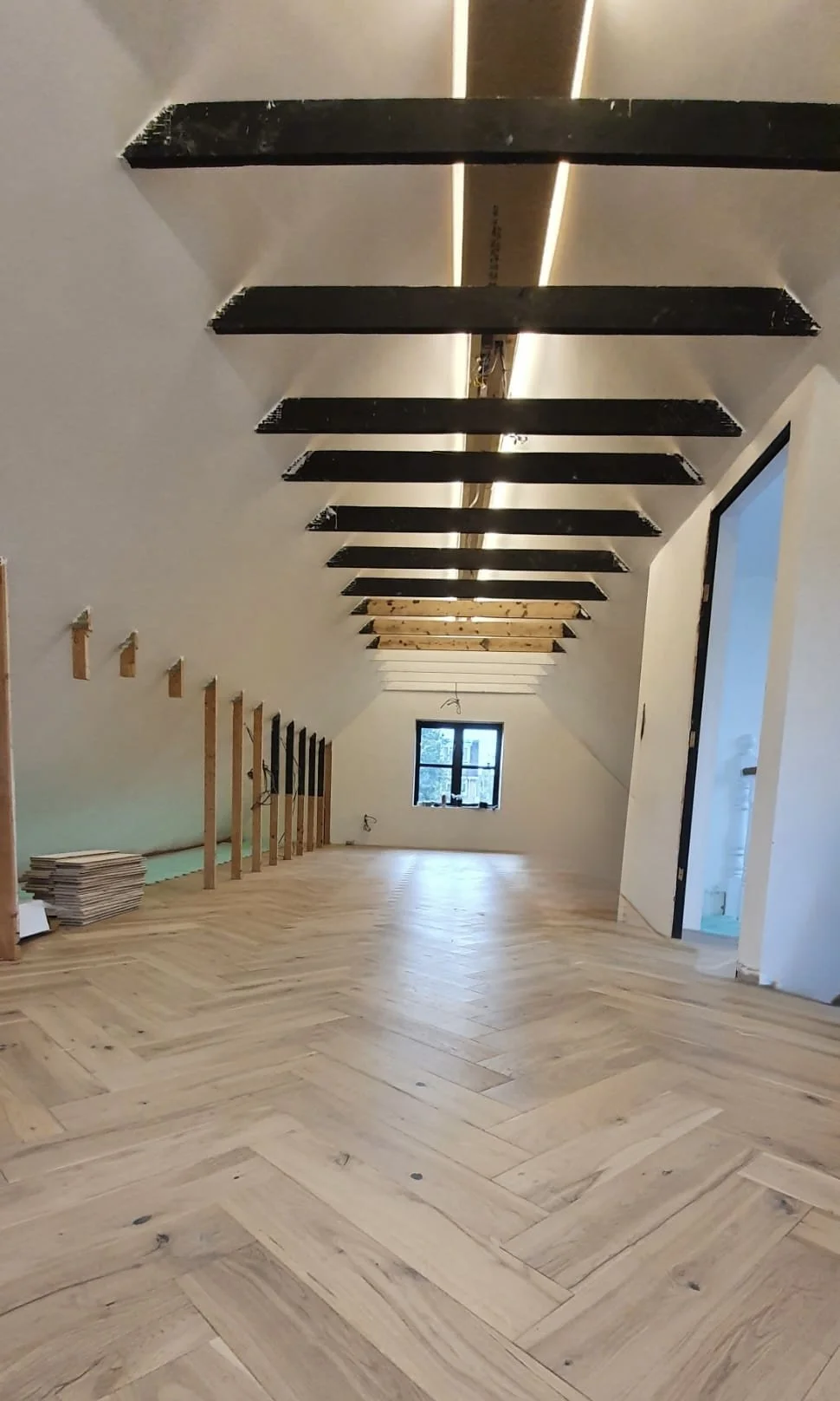 A spacious attic room with wood flooring, exposed ceiling beams, and a small window at the end of the room. Part of the wall on the right shows an open door leading to another room, and on the left, vertical wooden support beams are visible with some
