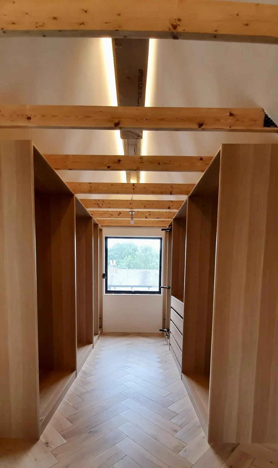 Partially constructed walk-in closet with wooden shelves and a window at the end, visible wooden beams on the ceiling, and hardwood flooring.