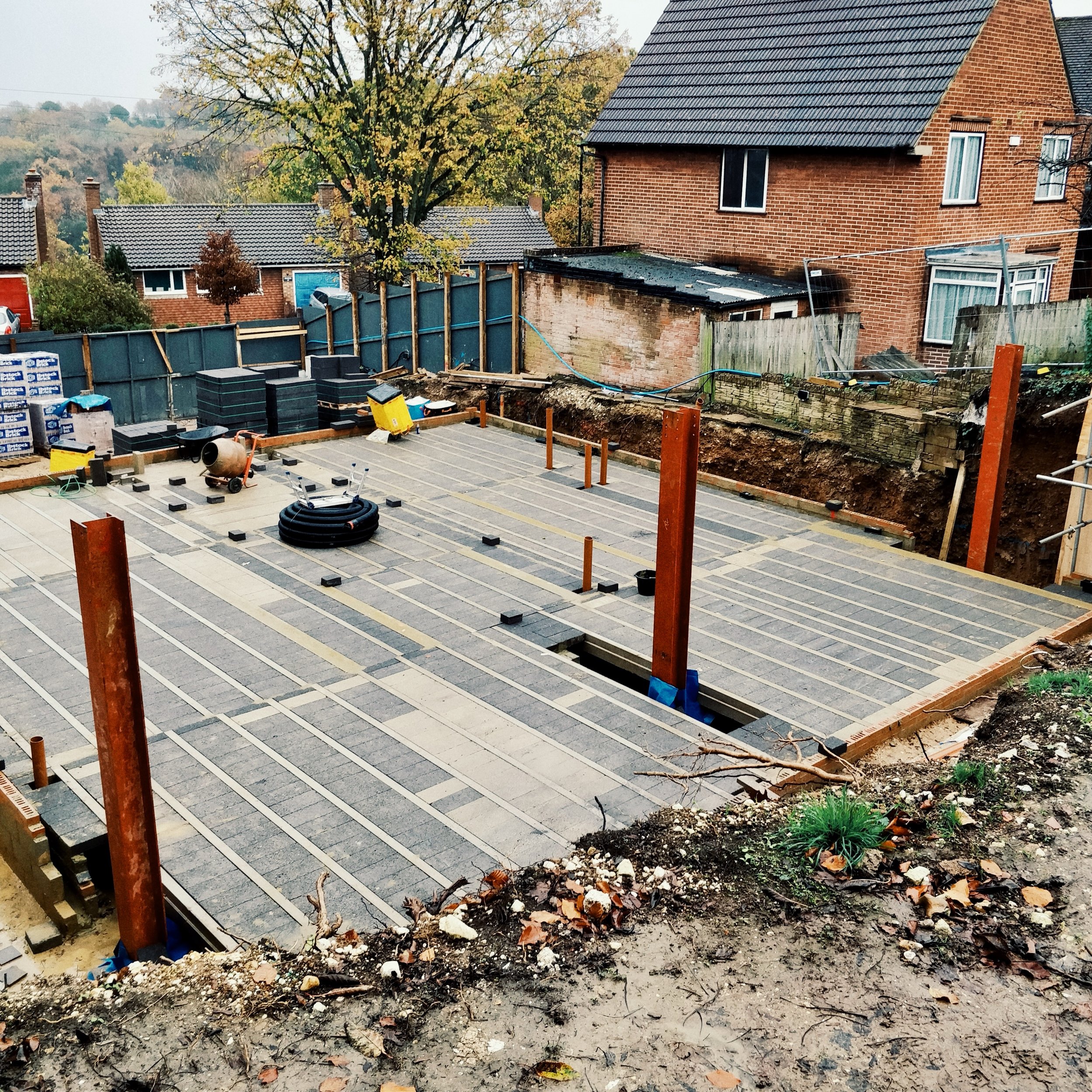 Construction site with a concrete foundation, wooden posts, construction materials, and surrounding residential houses with trees in the background.