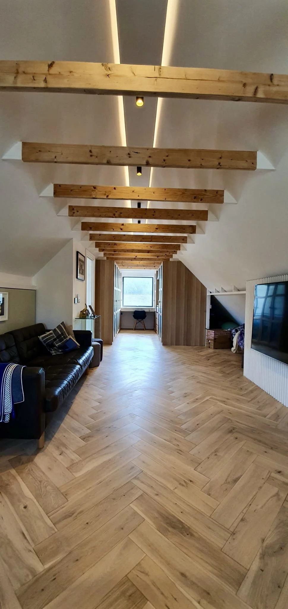 A spacious attic room with wooden beams on the ceiling, hardwood herringbone flooring, a black leather sofa with pillows on the left, a small table with a decorative item, a window at the far end, and a TV on the right.