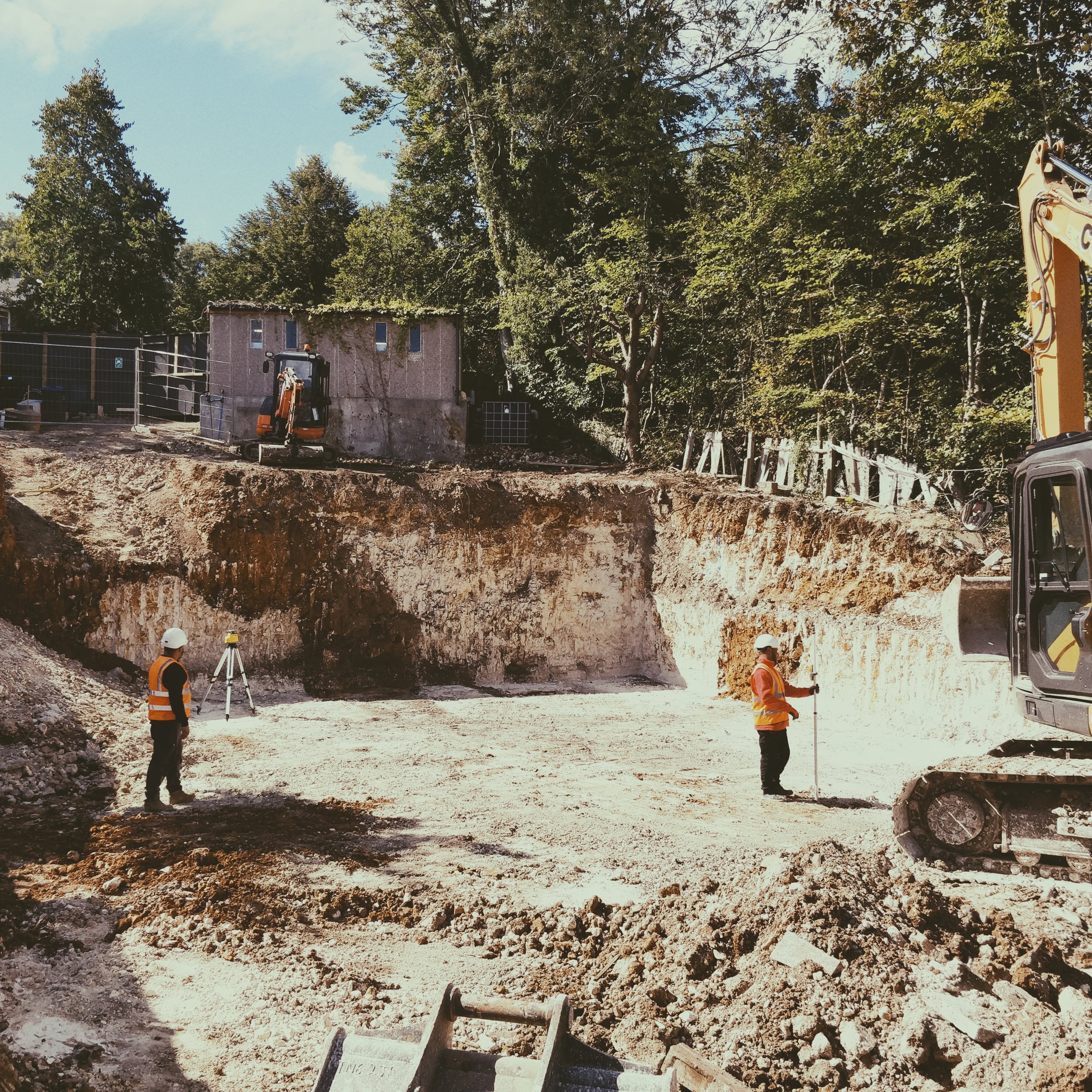 Construction site with workers in safety gear, excavator, and survey equipment, excavating a large hole in the ground surrounded by trees.