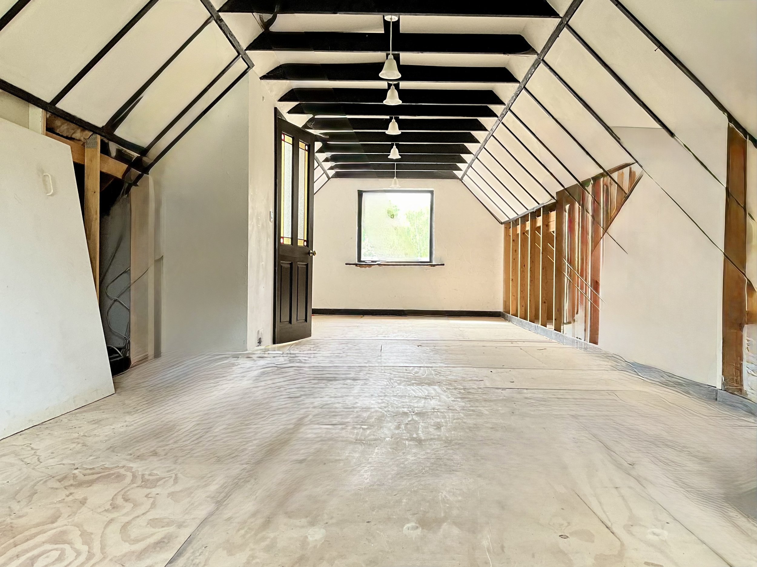 Interior of an unfinished room with exposed beams and framing, a window at the far end, and a partially constructed wall with electrical wiring, during construction or renovation.