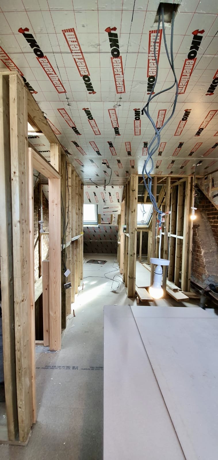 Interior of a building under renovation with exposed wooden framing, insulation, and electrical wiring. Ceiling has insulation panels marked 'IKO.'