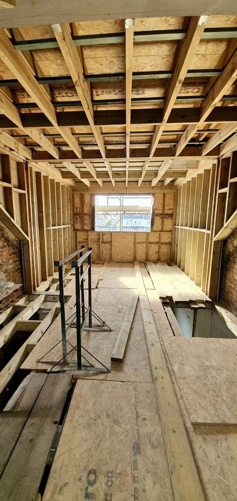 Interior view of a house under construction, showing wooden framing, an open window, and scaffolding.