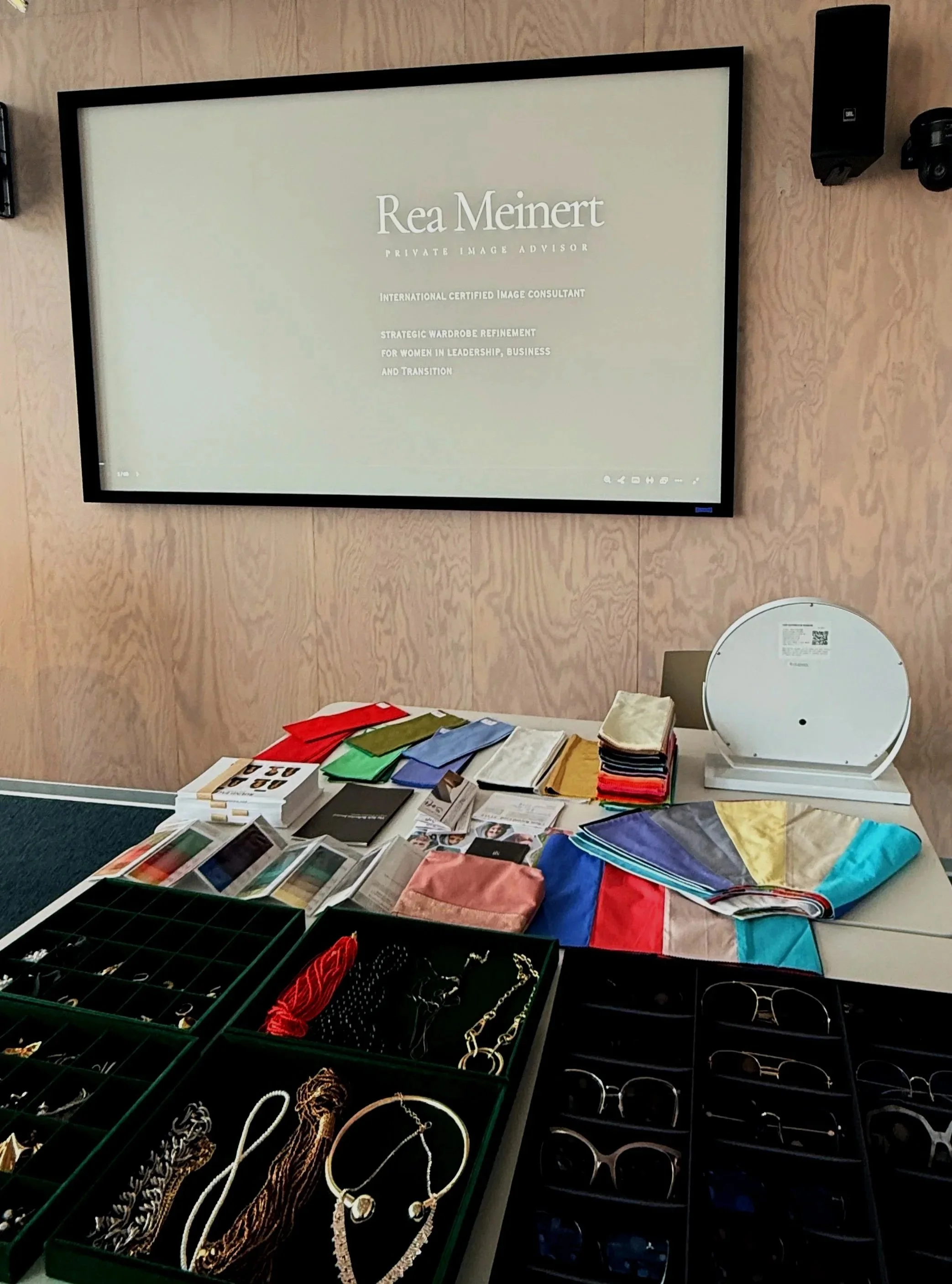 Display table of jewelry, including necklaces, bracelets, and earrings, with fabric swatches and color samples in the background. A large screen on the wall shows text about Rea Meinert, a private image advisor.