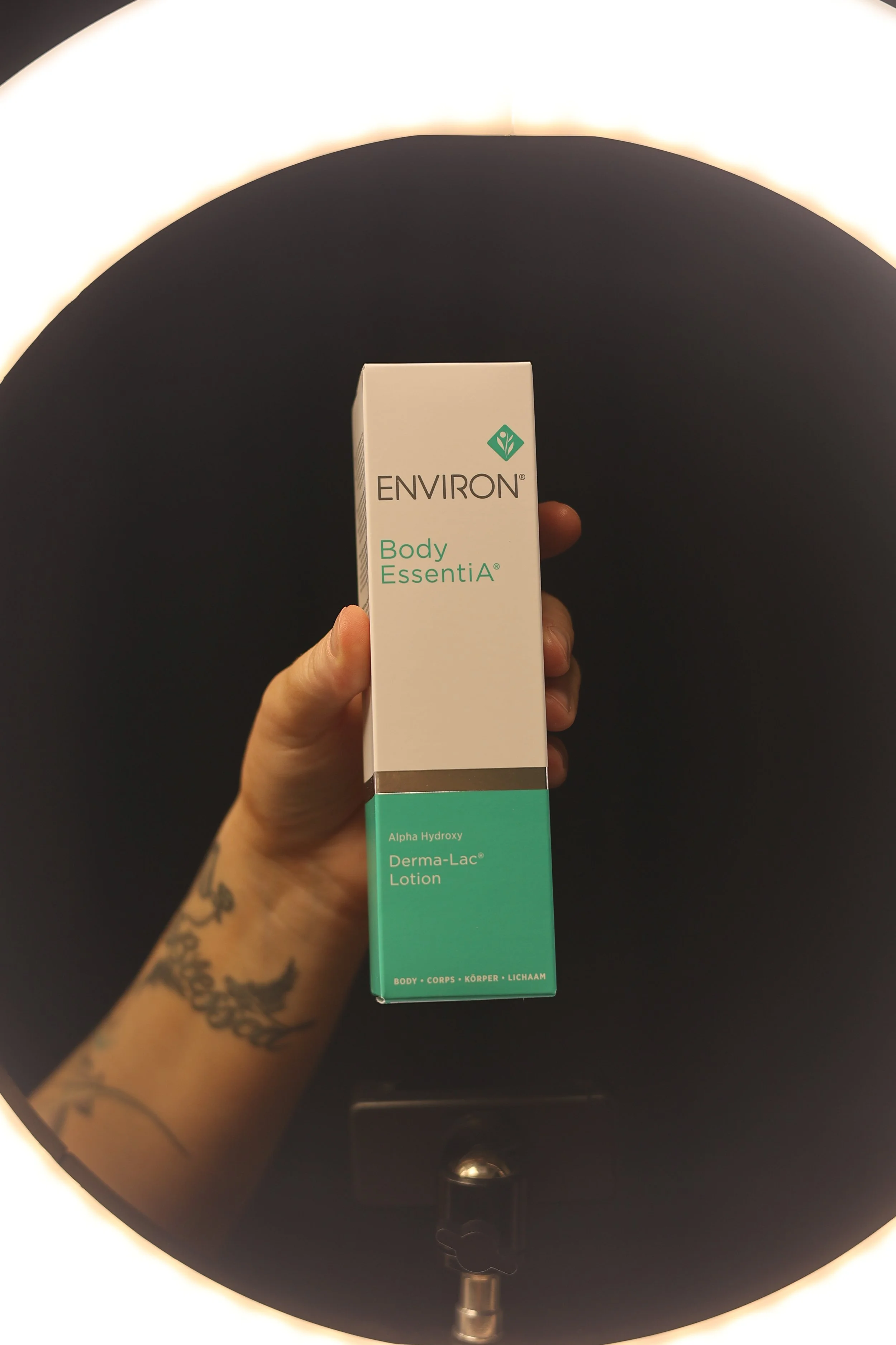 A hand holding a box of Environ Body EssentiA Derma-Lac Lotion pigment on a black circular surface with a bright light above.