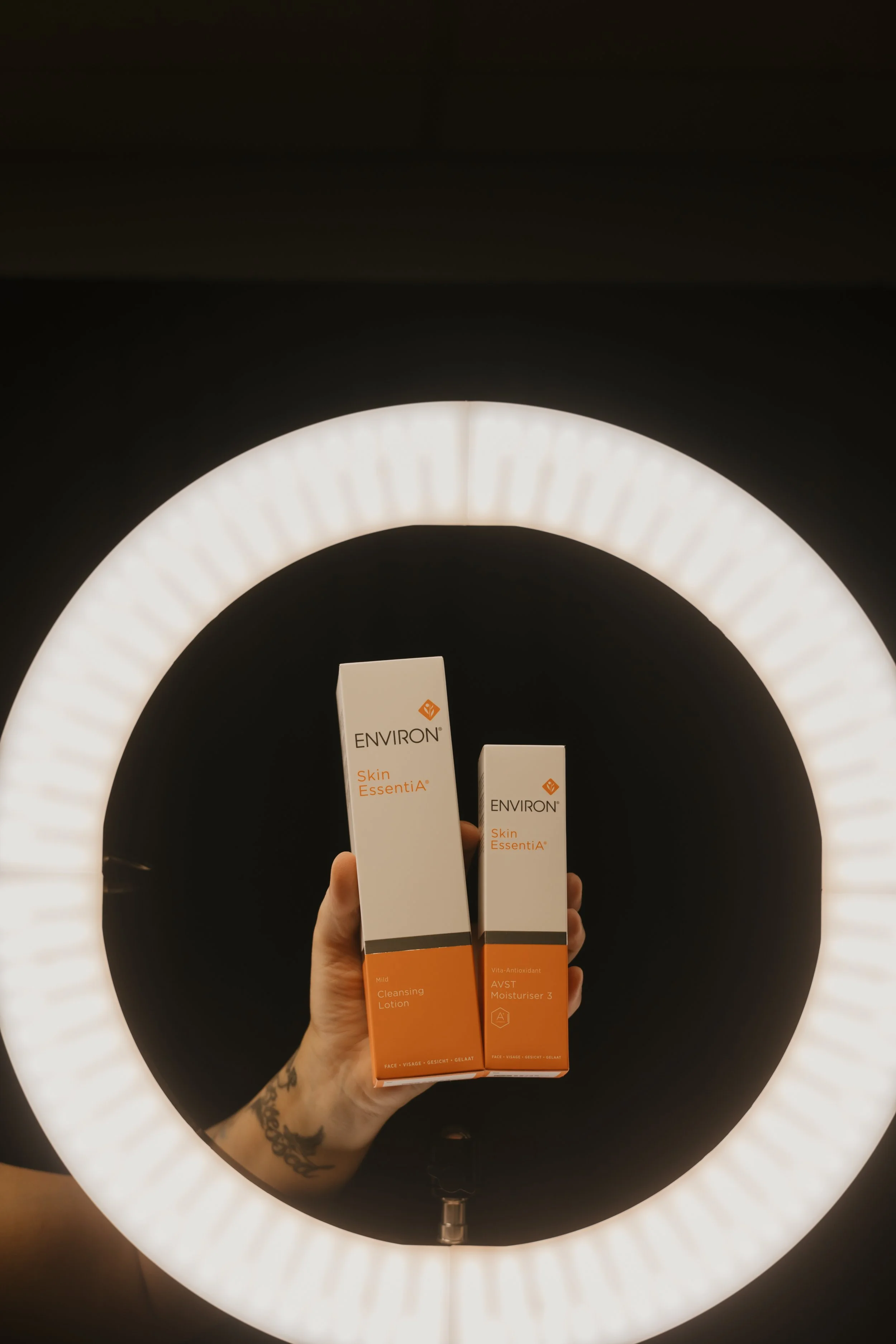 A person with a tattoo on their wrist holding two boxes of Environ Skin EssentiA products, a cleansing lotion and a moisturizer, in front of a large, circular, illuminated ring light against a dark background.