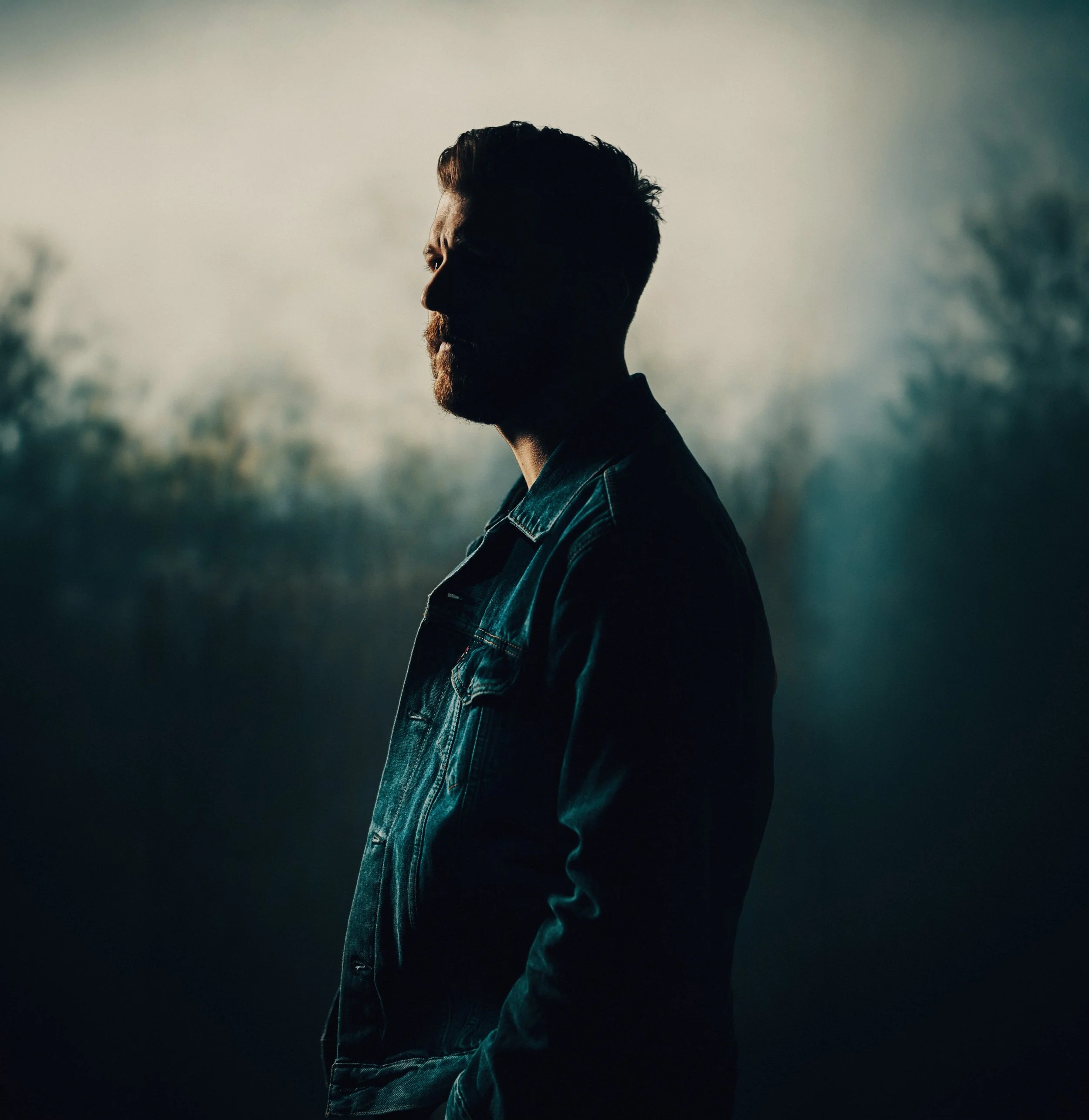 Side profile of a man with a beard and styled hair, wearing a denim jacket, standing outdoors during cloudy weather with blurred trees in the background.