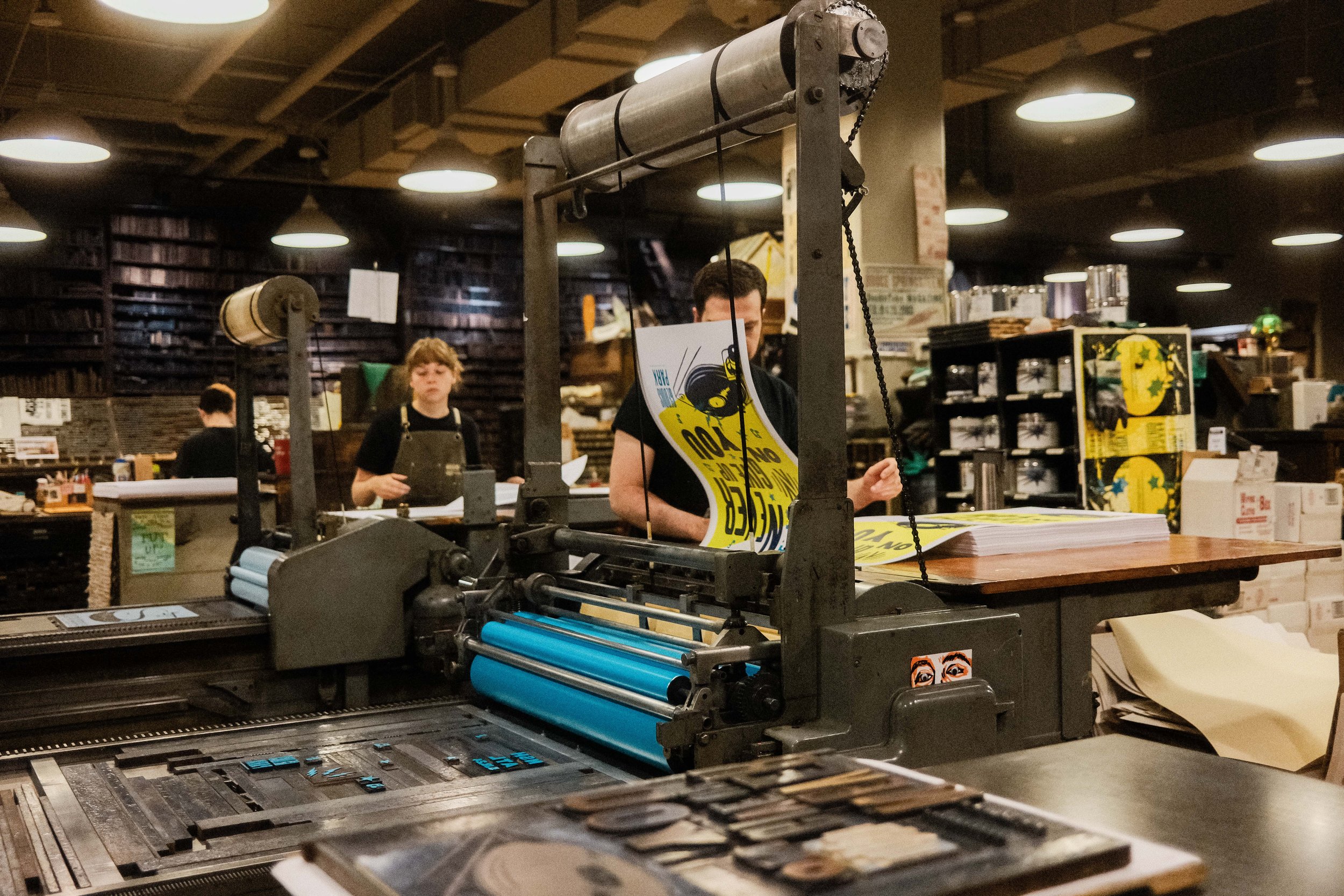 A printing press in a bookstore or print shop with people working in the background.