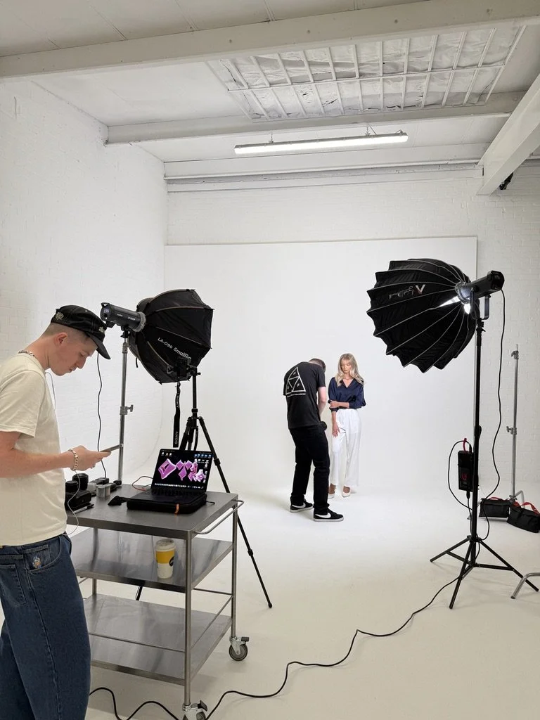 Photo shows a photo shoot setup in a white studio with three people. One person is on the left checking their phone, while a photographer and an assistant are working with a model dressed in a blue top and white pants.