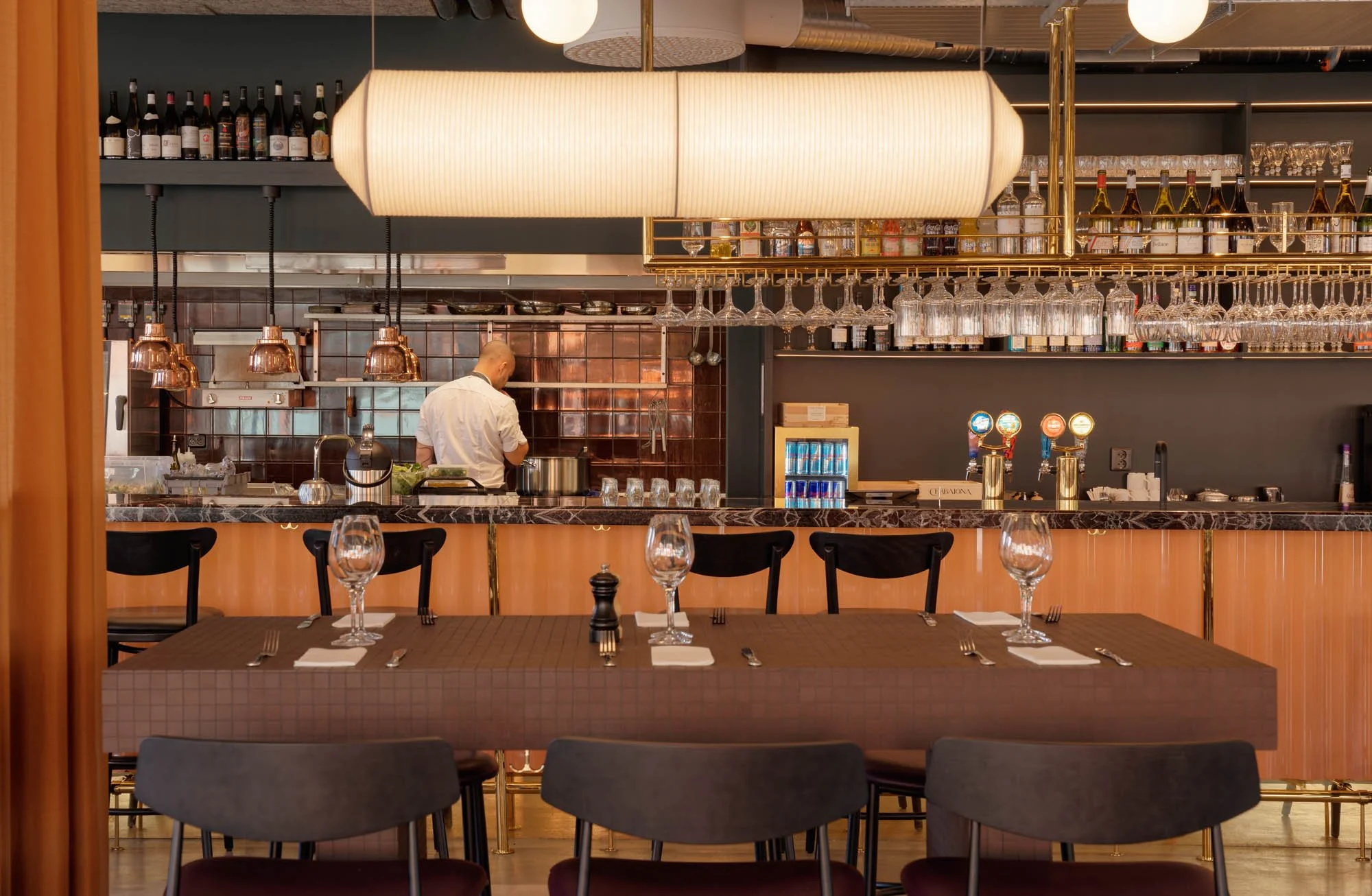 A view of a restaurant interior showing a bar area with hanging wine glasses, bottles of wine, and a bartender preparing a drink. Dining tables with place settings are in the foreground.