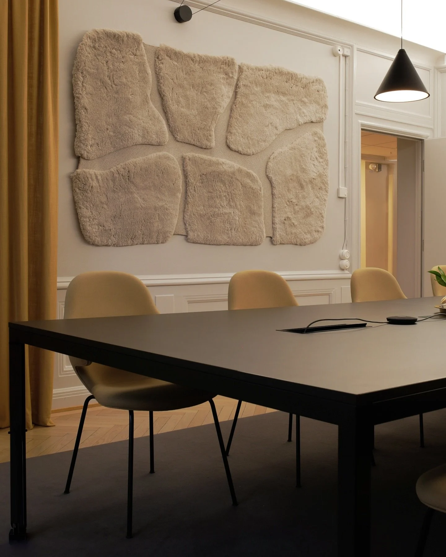 A meeting room with a black conference table and beige chairs, a large wall art installation of textured beige rocks, a black pendant light, and a doorway leading to a hallway.