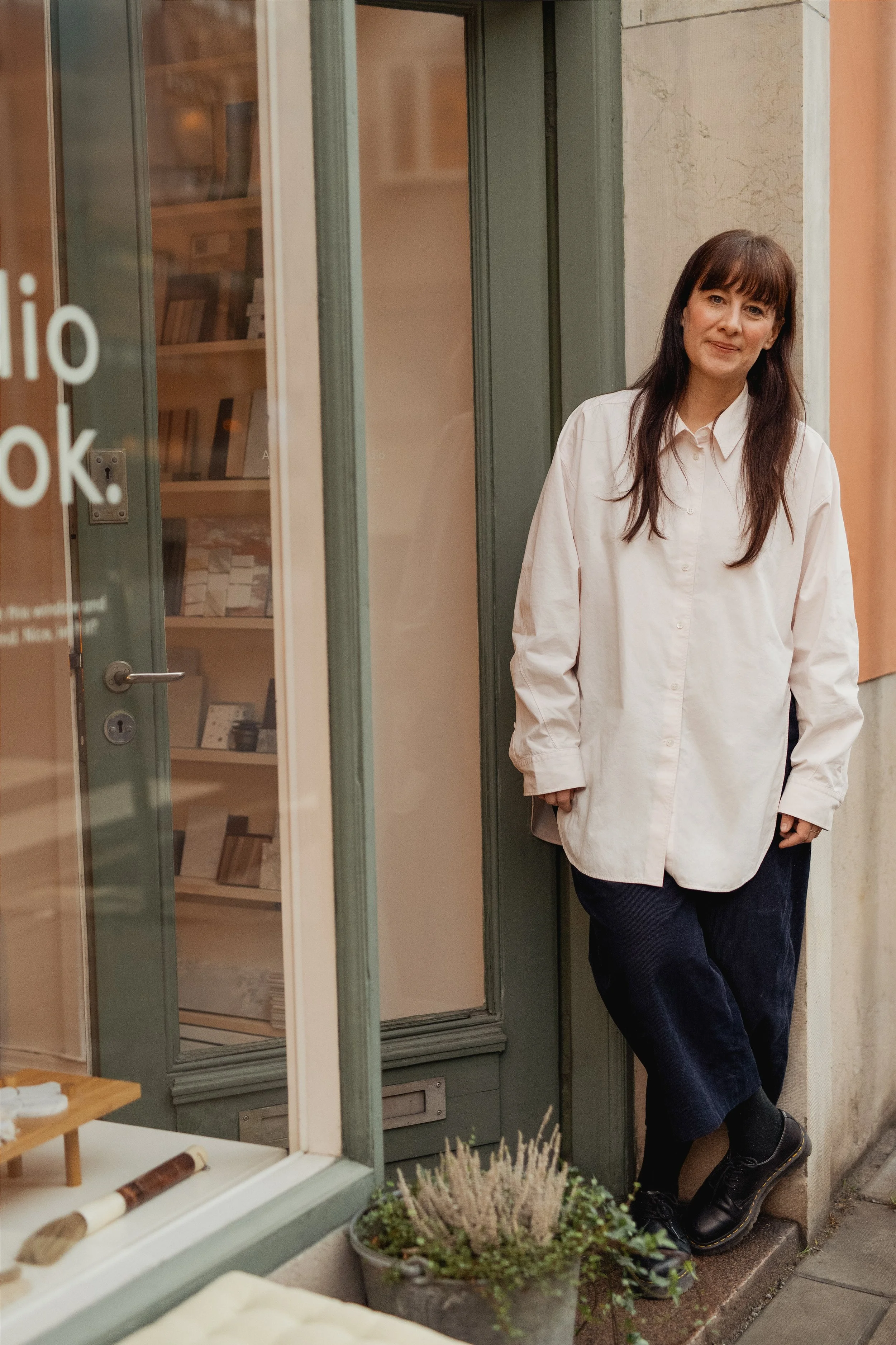A woman with long dark hair wearing a white oversized shirt and dark pants, standing at a doorway with a slight smile, against the backdrop of an outdoor building and small potted plants.