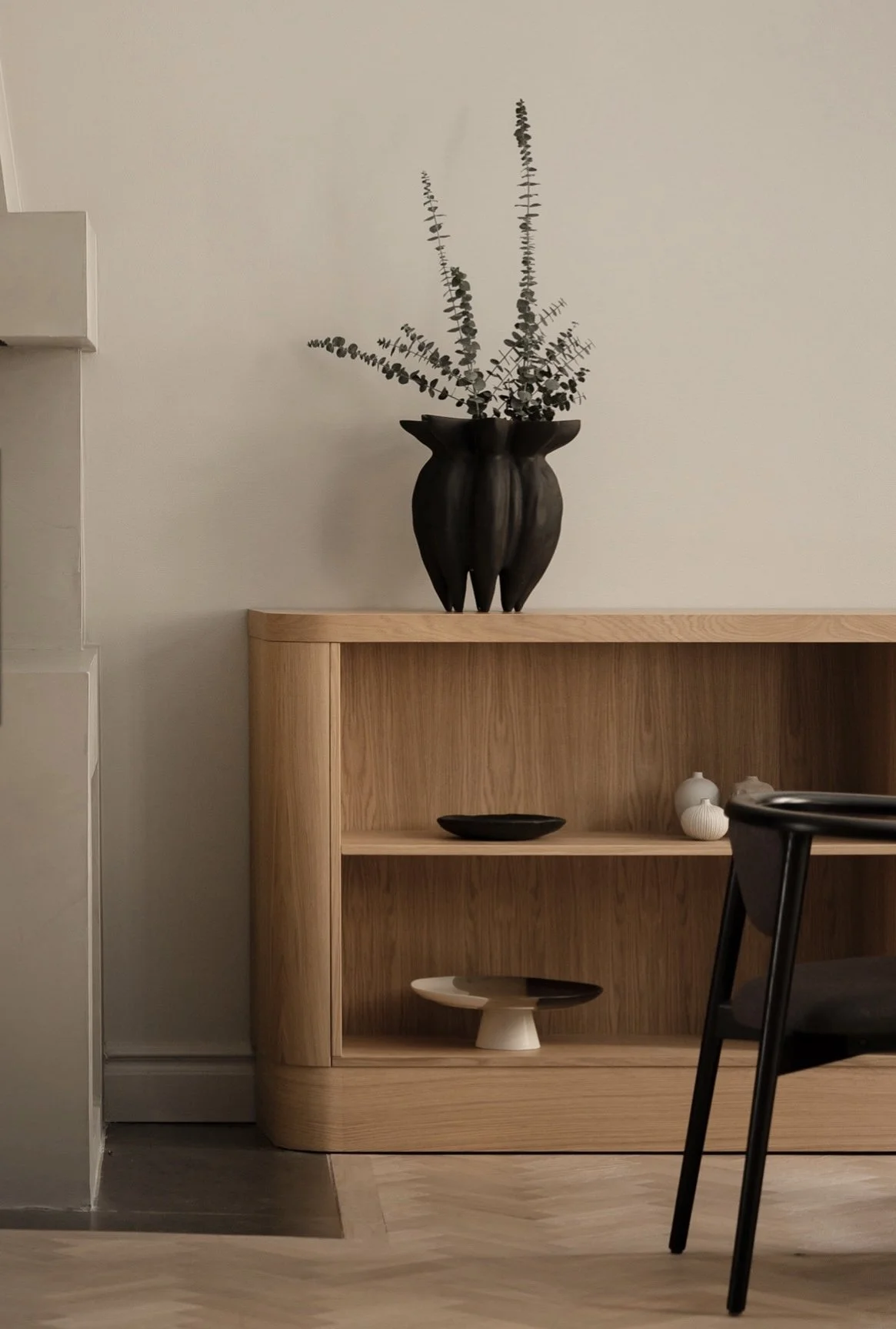 Wooden cabinet with decorative vases and bowls, large black vase with eucalyptus branches on top, black chair partially visible in foreground.