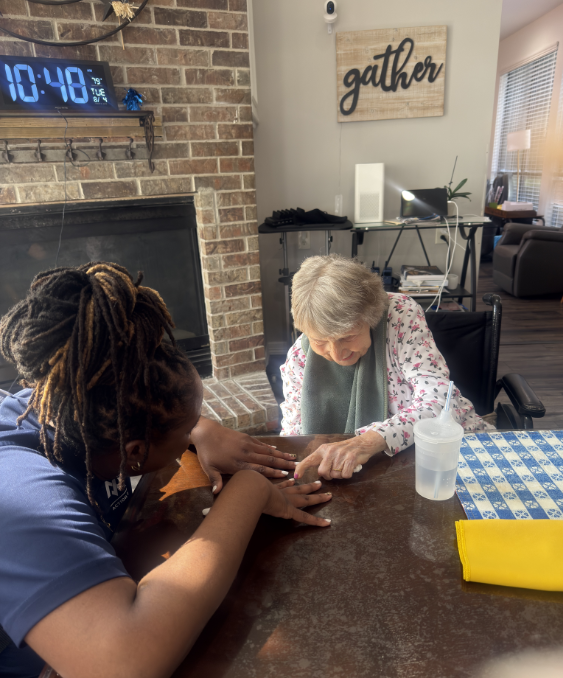 Care That Connects: Kim sitting hand-in-hand with a resident during a quiet one-on-one moment, offering the kind of gentle, personal attention that makes a house feel like home.