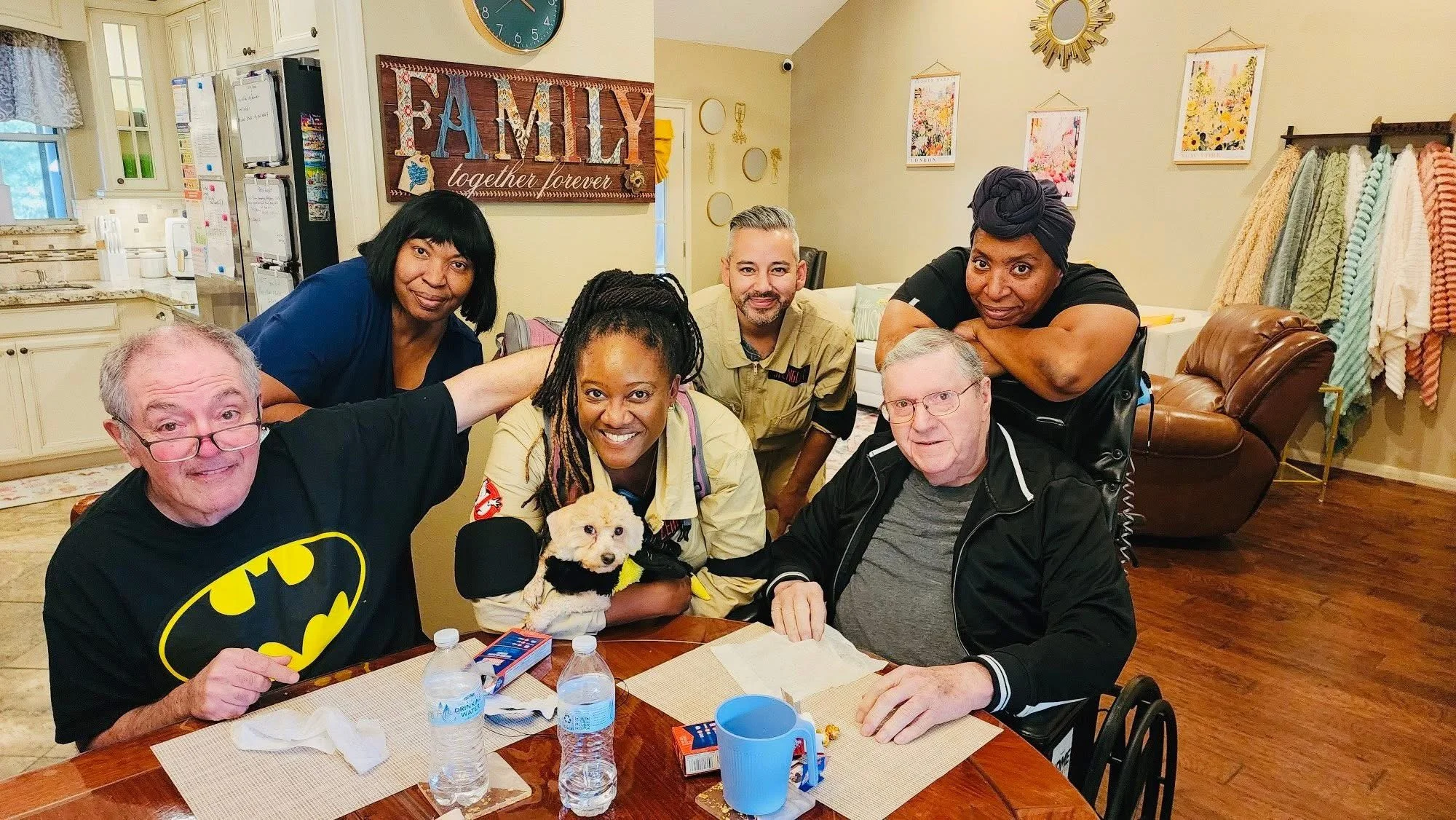 Chris and Kim Visiting an Assisted Living Facility: Chris and Kim are visiting a resident in an assisted living lounge, sharing a friendly, relaxed moment together while everyone smiles and the atmosphere feels welcoming.