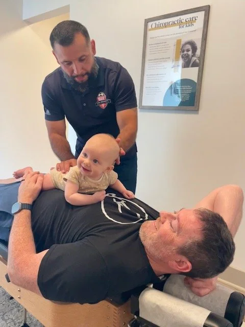 A chiropractor with a beard and a dark shirt adjusts a young girl lying on a chiropractic table. The girl is smiling and looking at the chiropractor. A man, presumably the girl's father, is lying on the table, smiling with his eyes closed. A framed poster about chiropractic care is on the wall behind them.